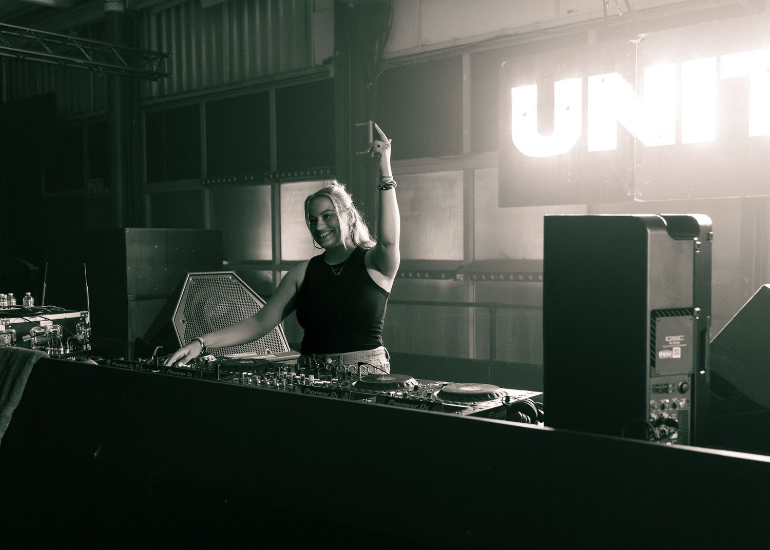 Female DJ performing at UNIT club, raising hand behind turntables in black and white photo. Festival photography,
