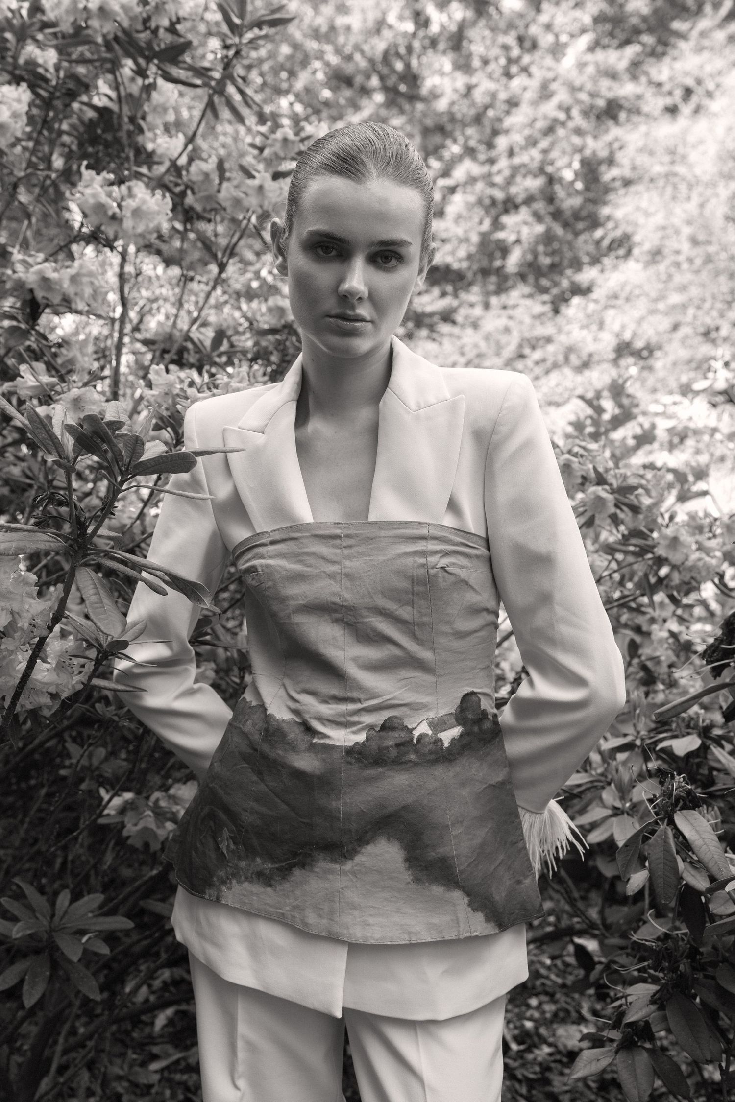 Black and white photo of a young woman in a white blazer and painted corset top, standing among lush garden foliage.  fashion photography,
