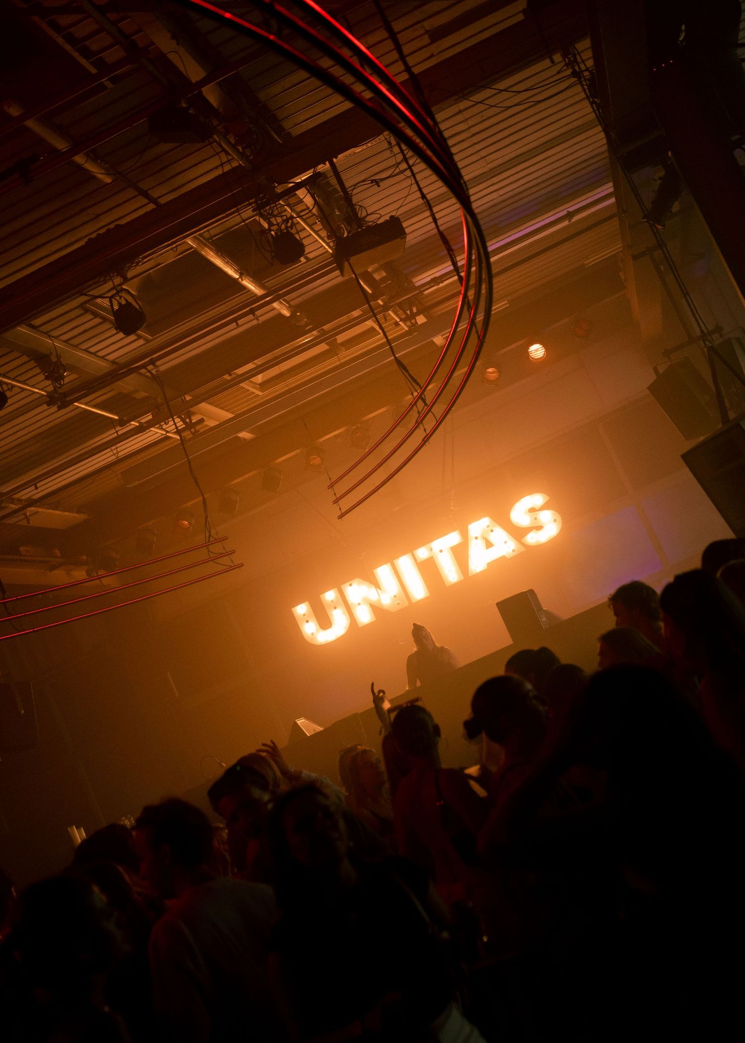 Crowd at Unitas nightclub event with glowing orange neon sign illuminating a dark industrial venue.