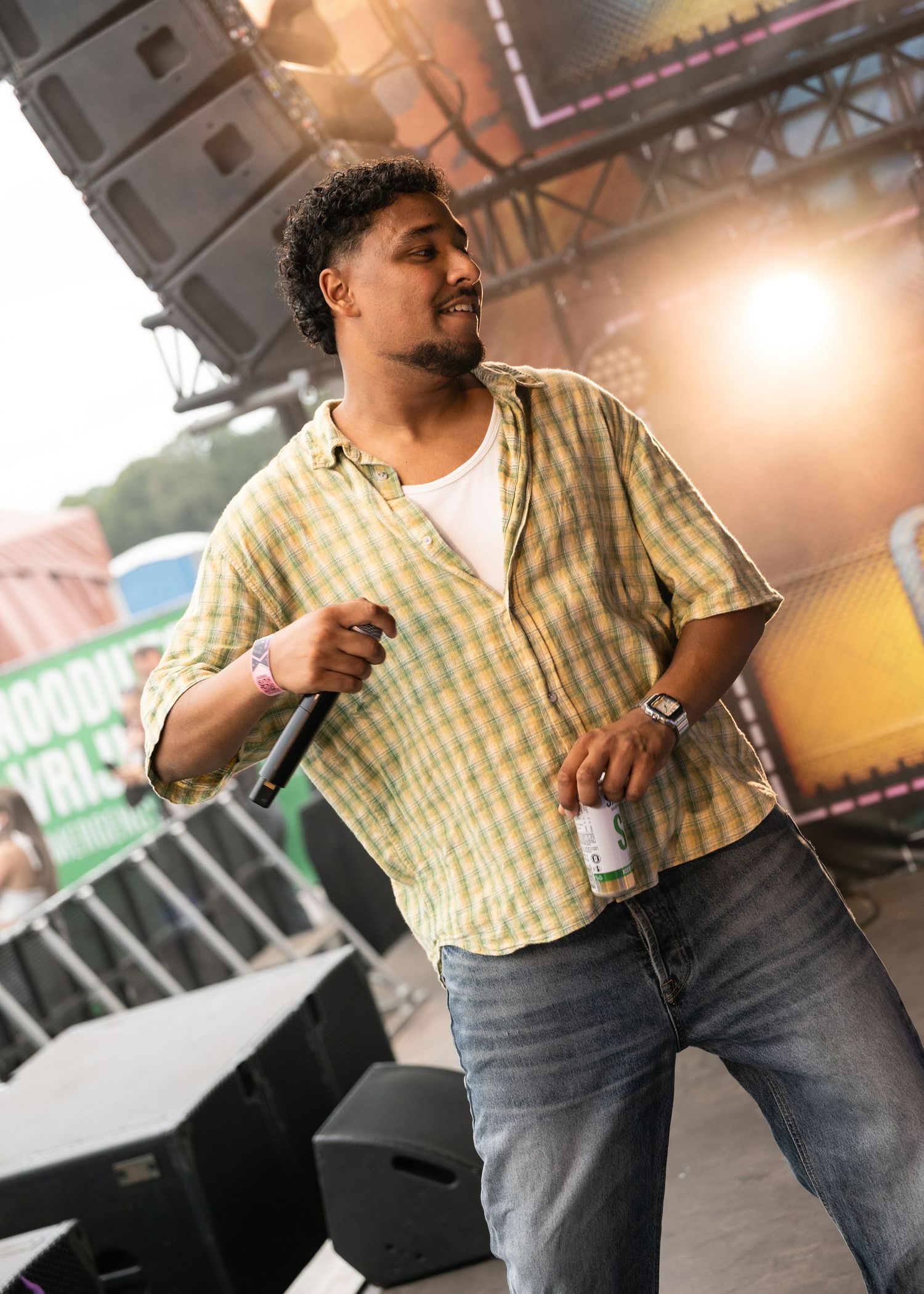 A man performs on stage holding a microphone and drink, wearing a green plaid shirt and jeans at an outdoor music festival.