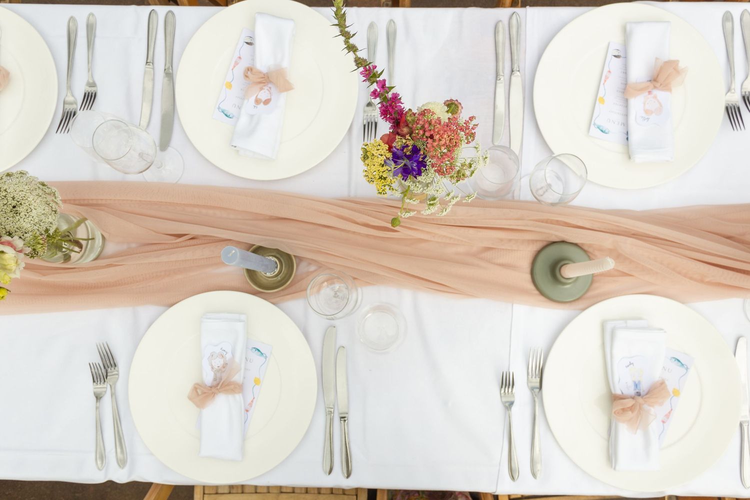 Elegant wedding table setting with blush runner, white plates, silver cutlery, and colorful floral arrangements viewed from above.