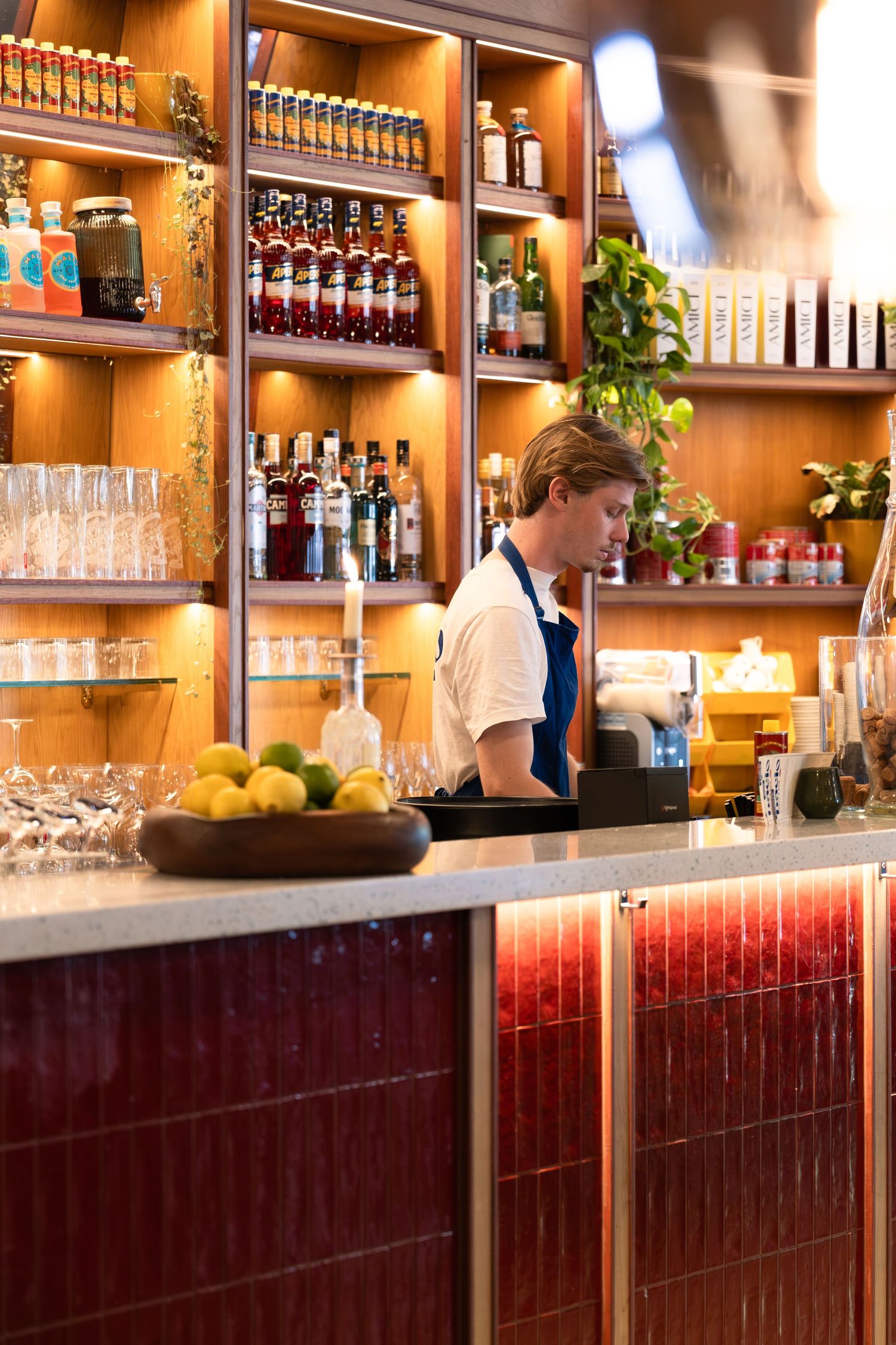 Bartender in blue apron working behind a warmly lit bar with shelves of spirits and glassware.