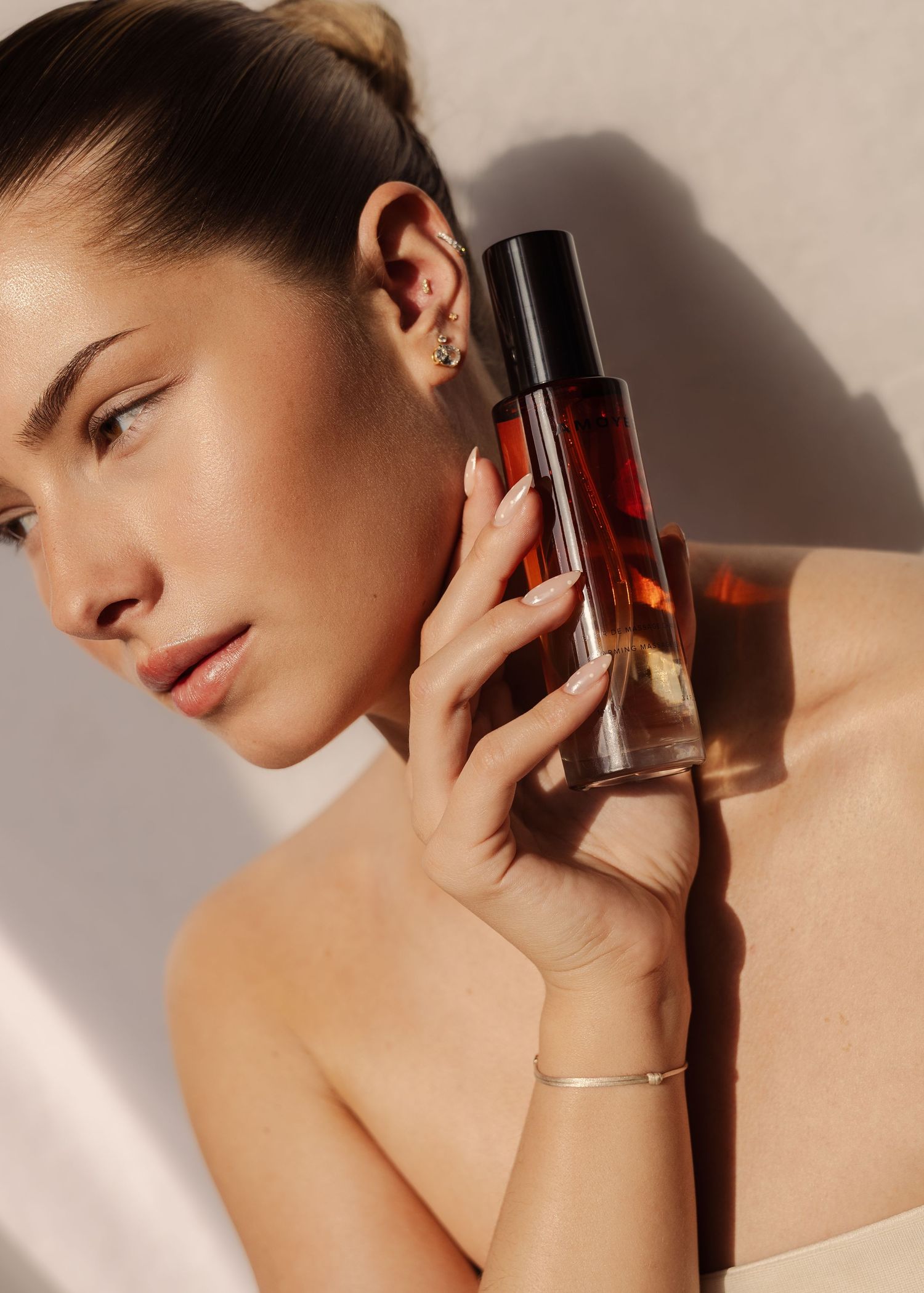 Woman holding oil bottle near her face in soft natural light, wearing gold earrings and a silver bracelet.   fashion photography, netherland