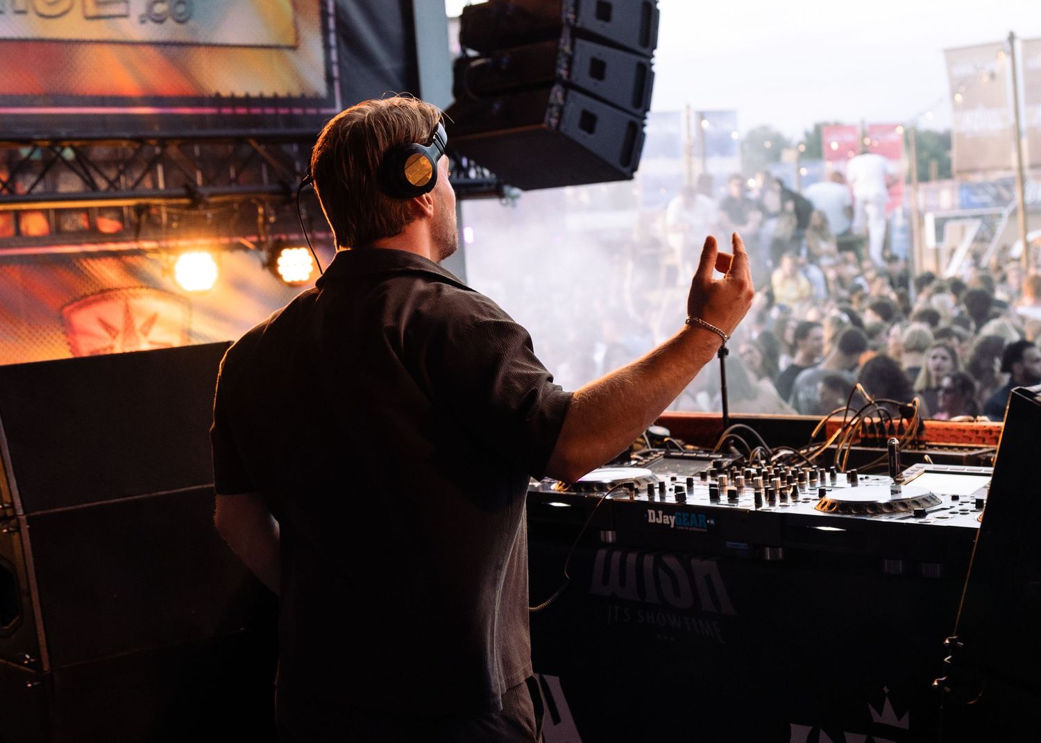 A DJ wearing headphones performs at an outdoor music festival, facing a large crowd with DJ equipment in the foreground. Festival photograph
