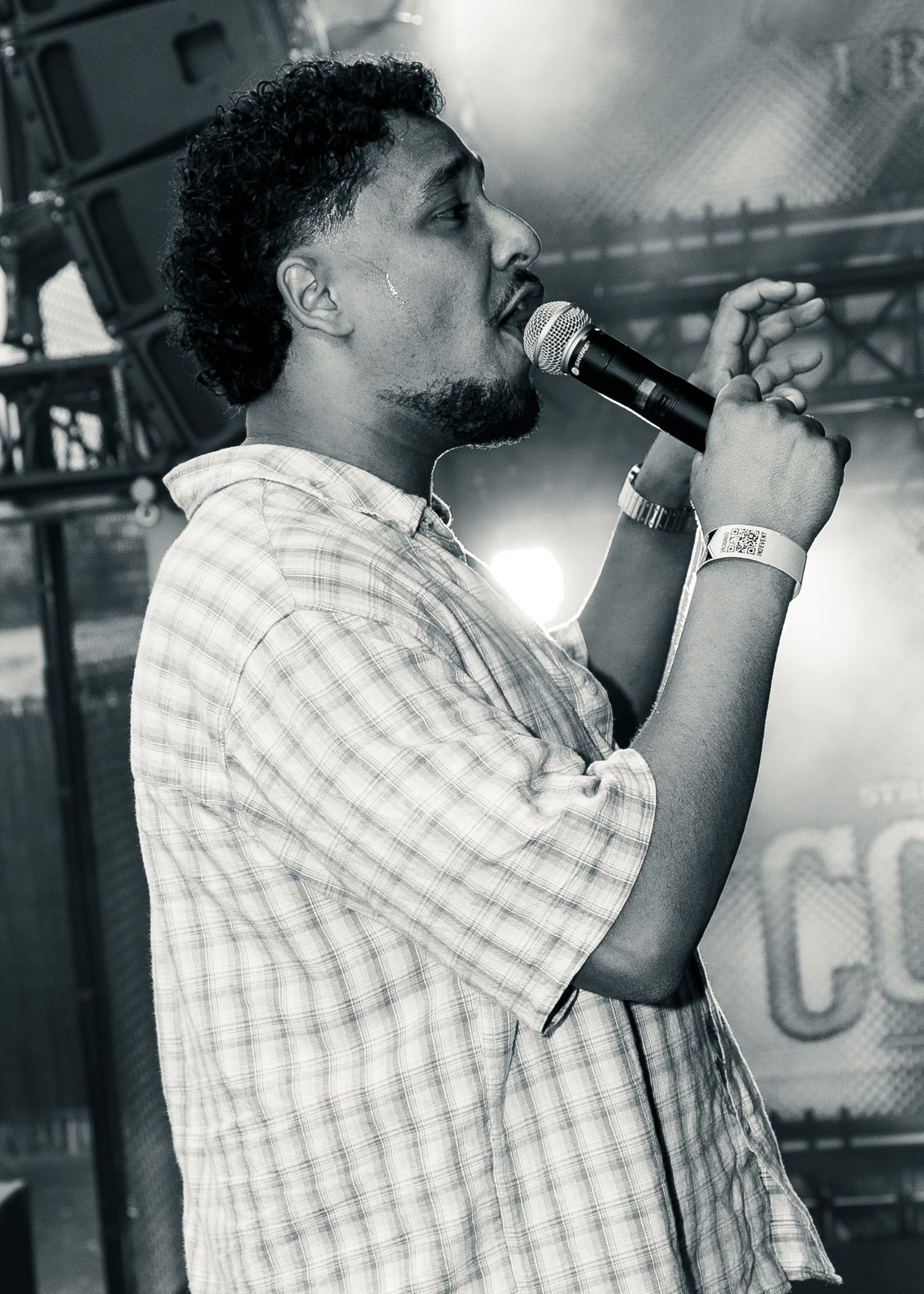 Black and white photo of a man singing into a microphone on stage, wearing a plaid shirt and watch. Festival photography,