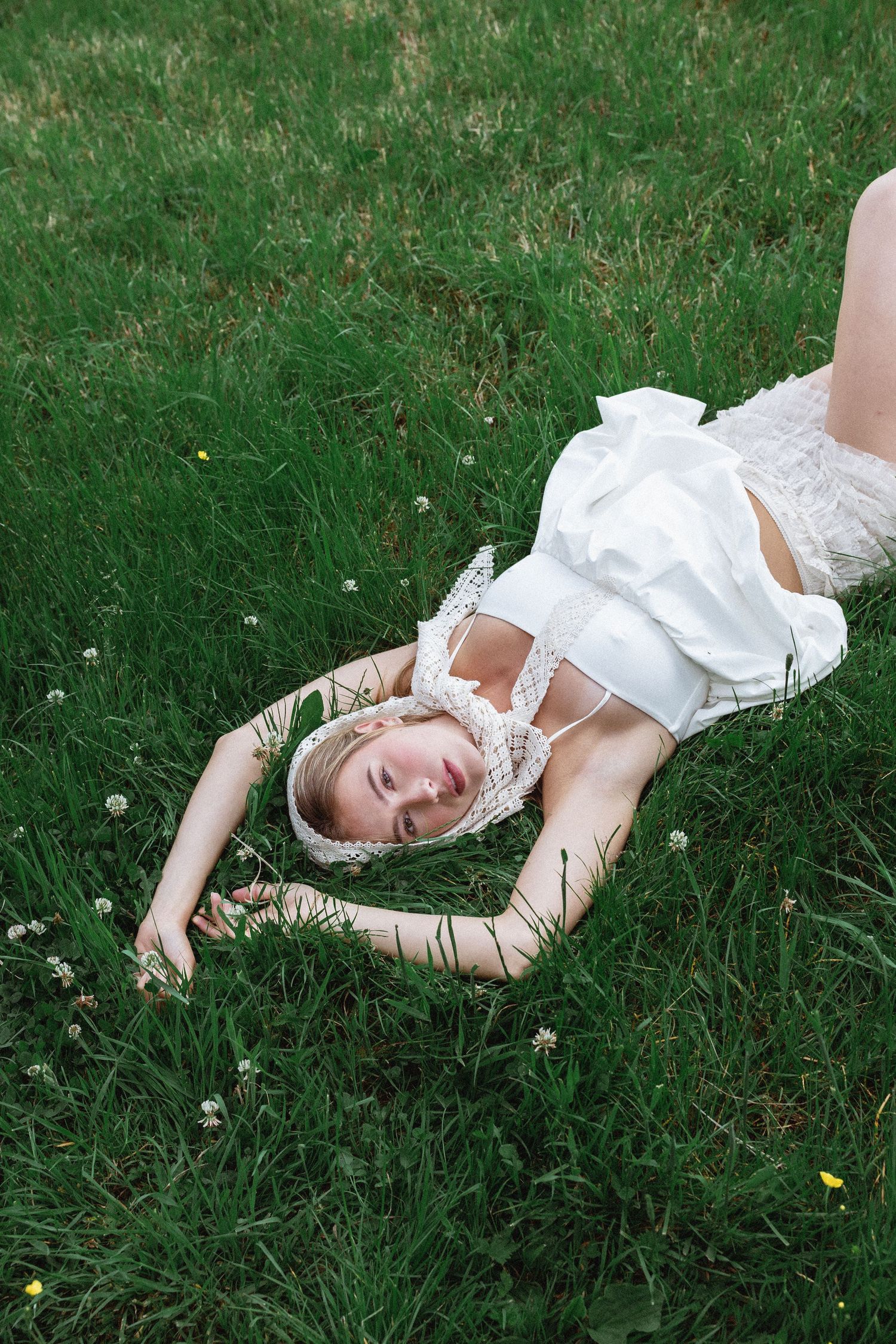 A blonde woman in a white dress lies peacefully on lush green grass with small wildflowers around her.  fashion photography,