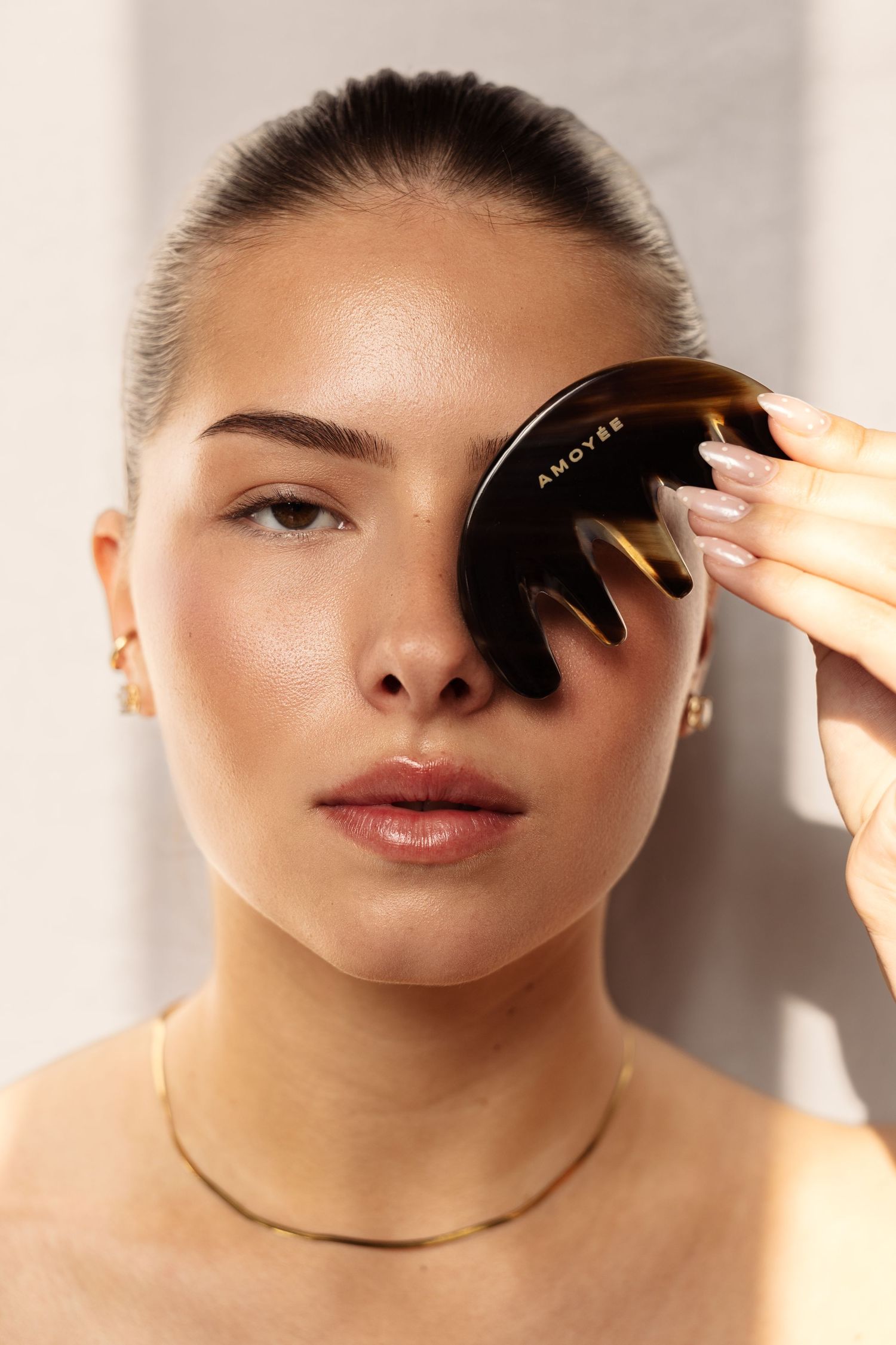 Woman holding a dark tortoiseshell Amoyre gua sha tool over her eye, wearing gold jewelry.  fashion photography, netherlands