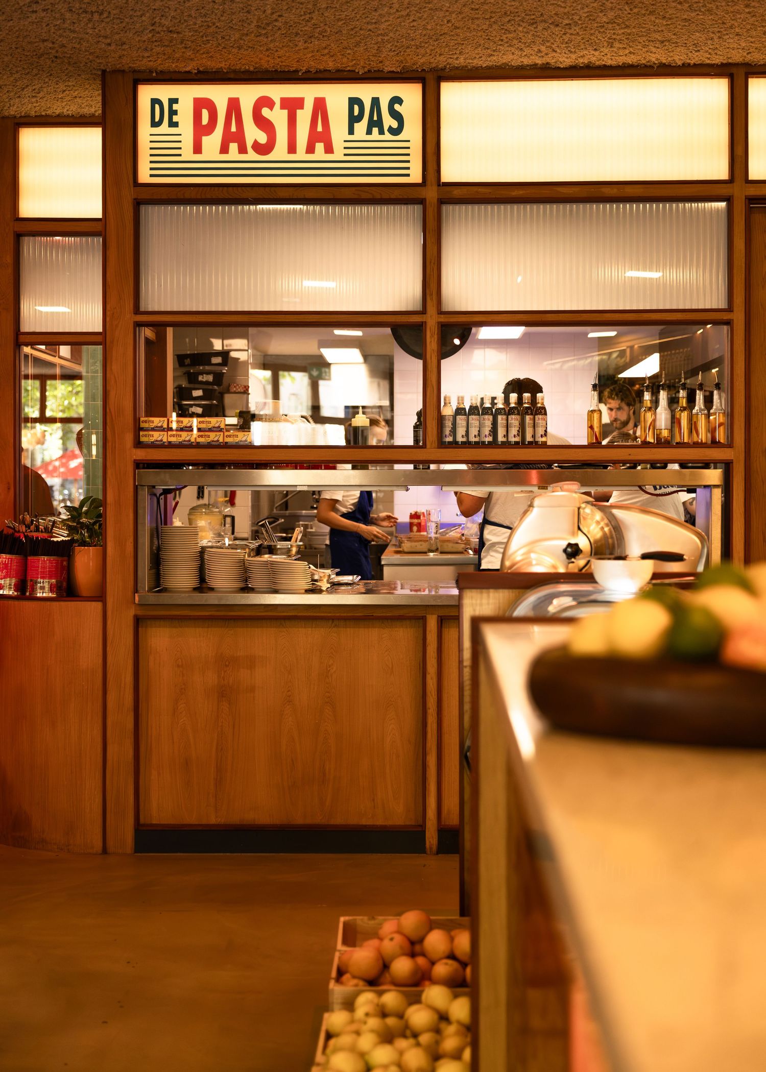 Interior of De Pasta Pas restaurant showing a warm wooden counter, open kitchen with chef, and fresh produce display.