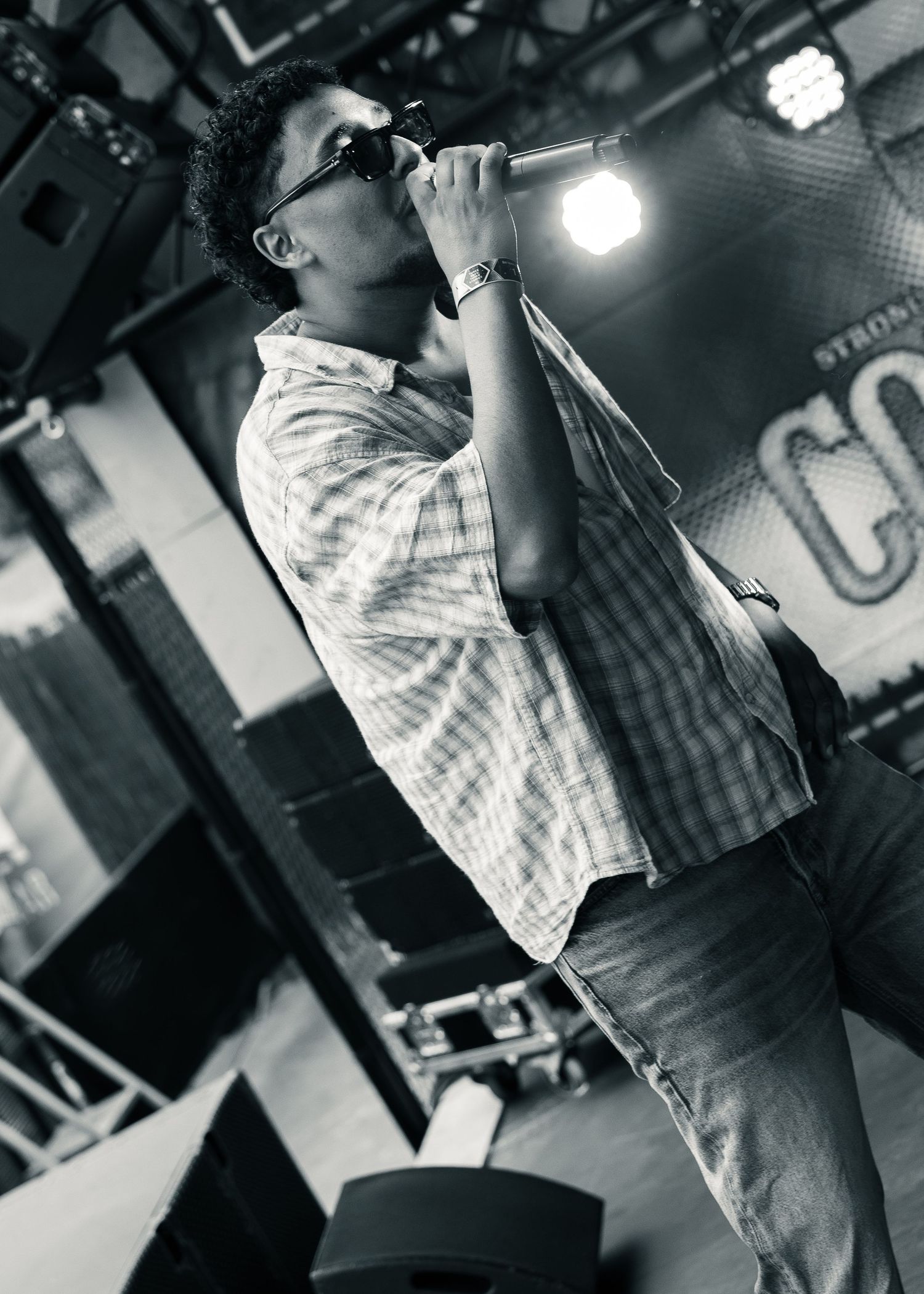 Black and white photo of a young man singing into a microphone on stage wearing sunglasses and a plaid shirt. Festival photography,