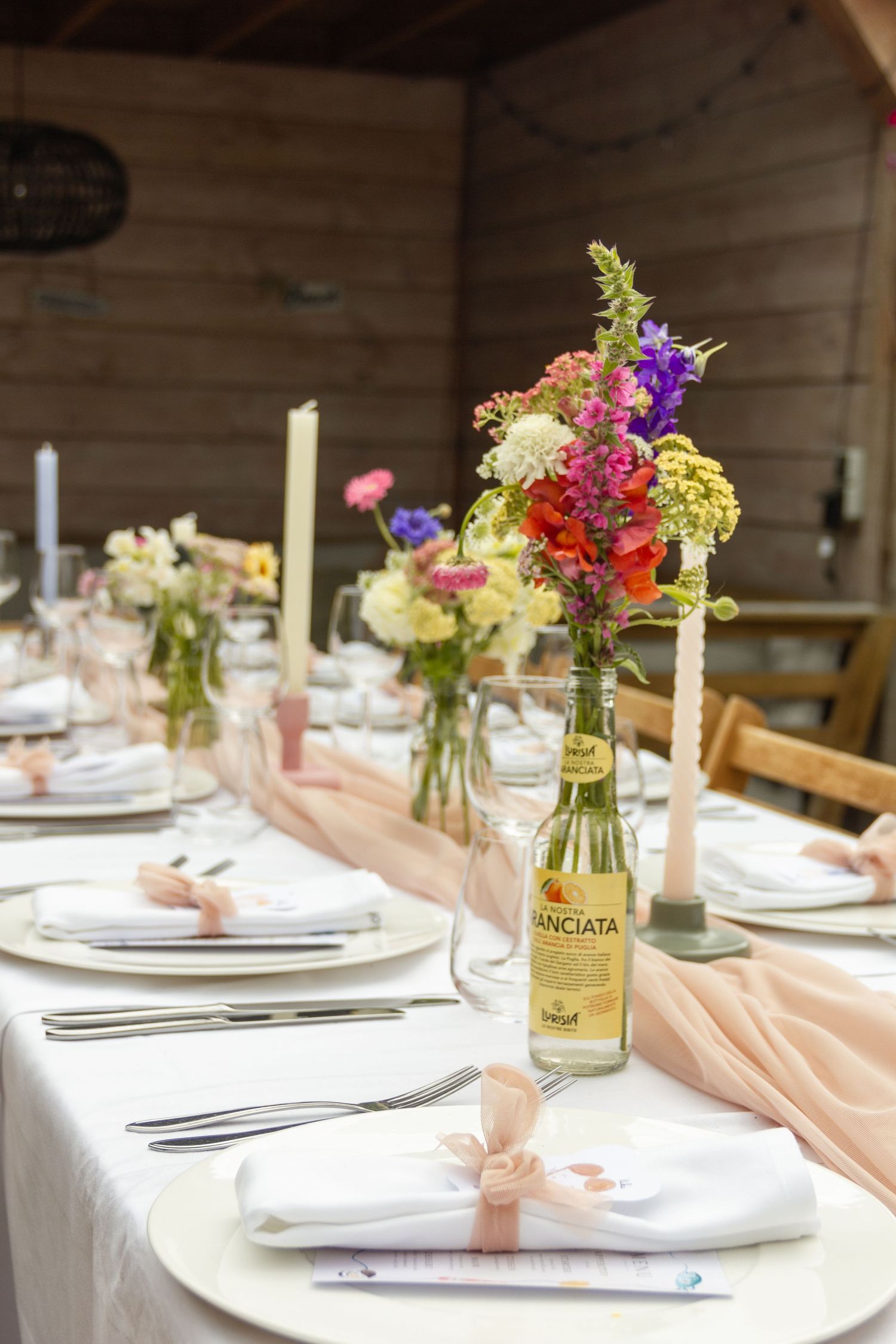 Elegant wedding table setting with colorful wildflowers in glass bottles, pink ribbon napkins, candles, and a Sanciata bottle centerpiece.