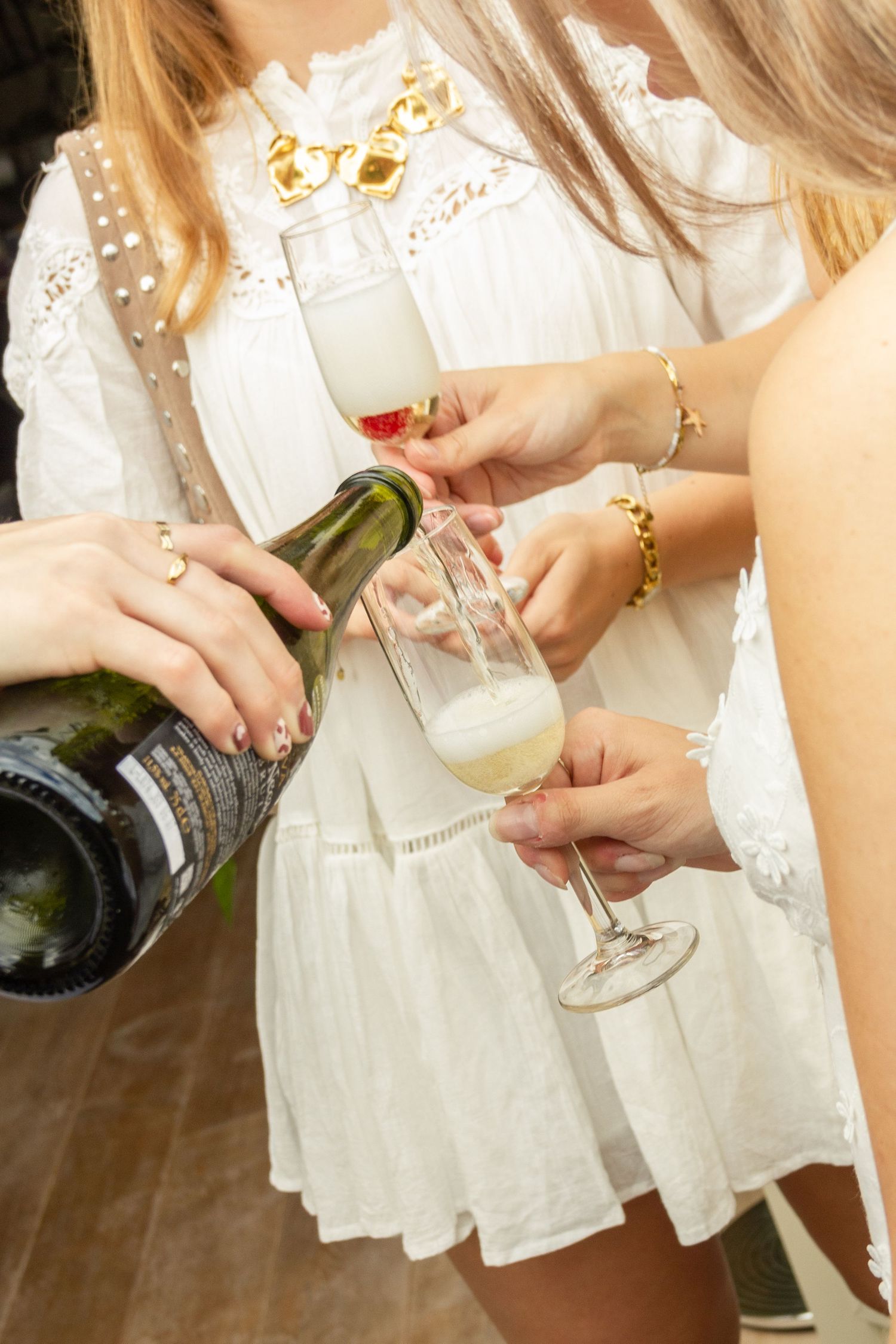 Women in white dresses celebrating with champagne glasses being filled from a green bottle at a party. event photography, netherlands