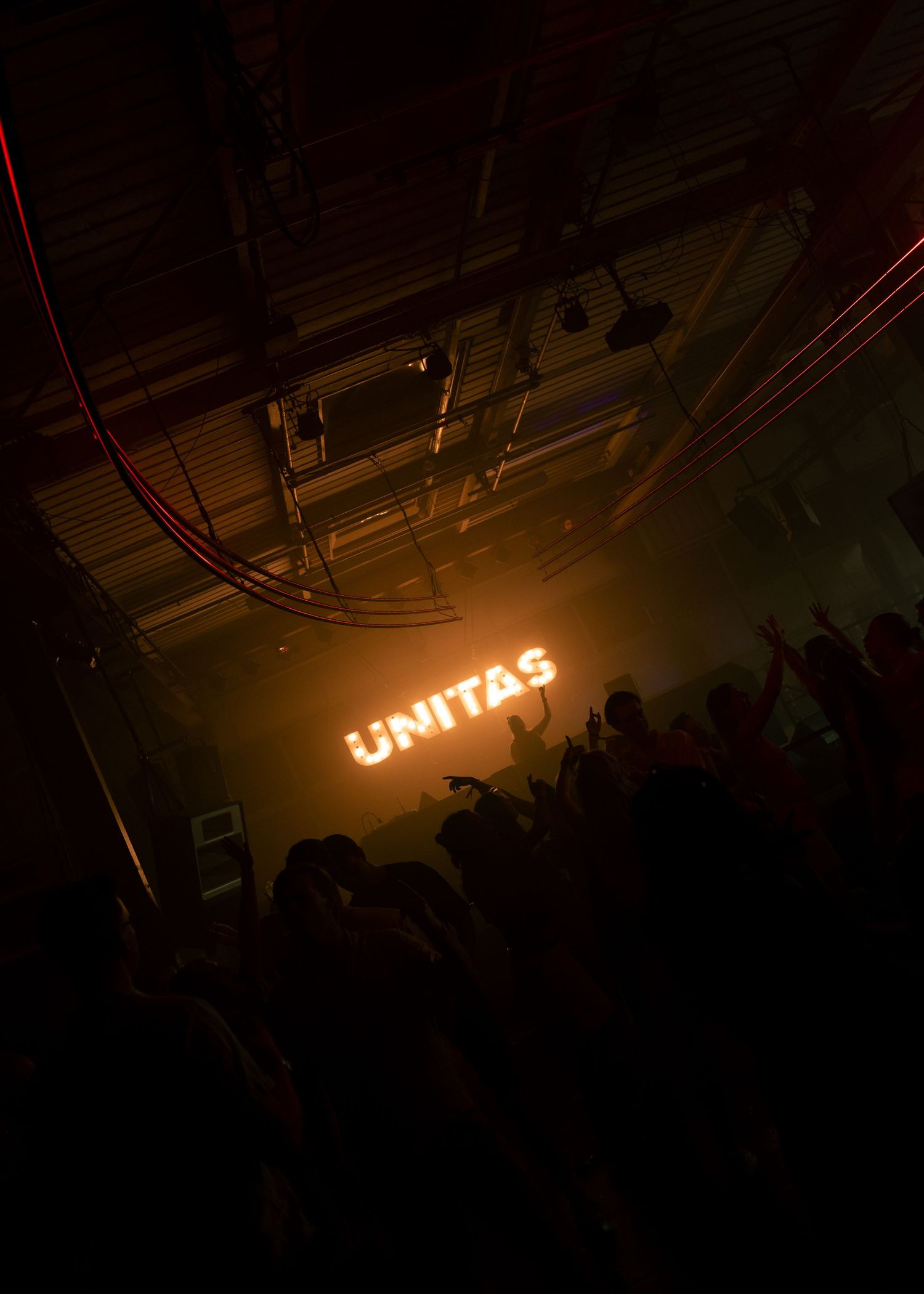Crowd at UNITAS nightclub event with glowing orange neon sign illuminating dark venue filled with raised hands. Festival photography,