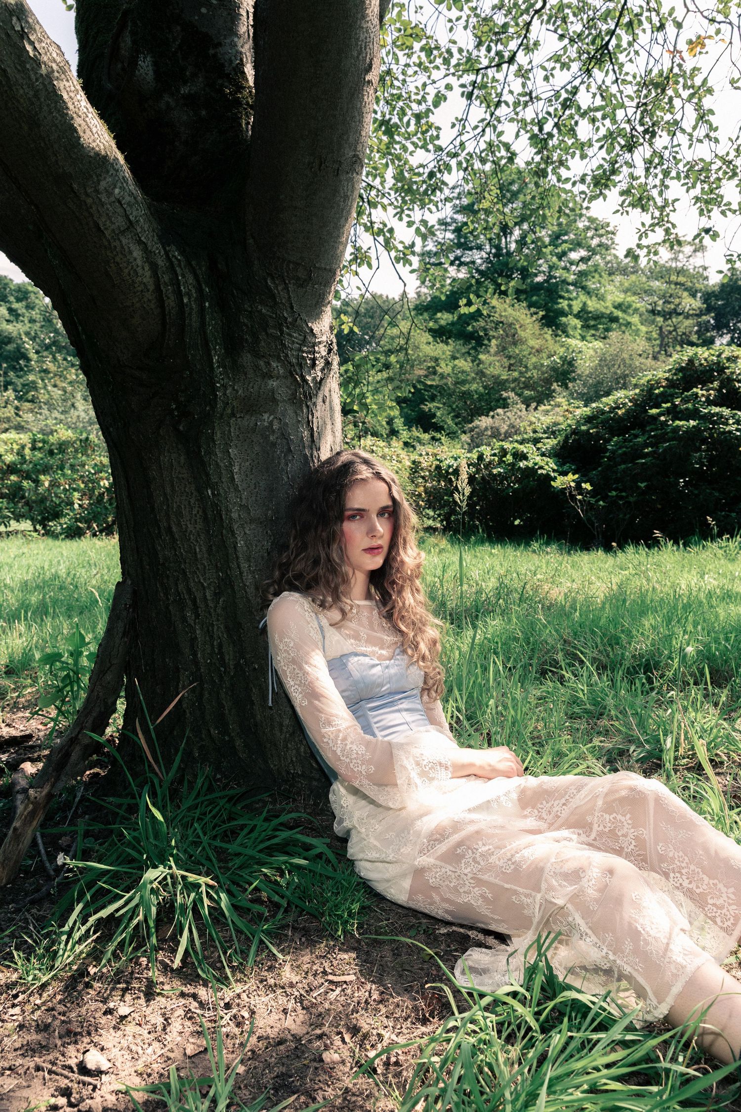Young woman in lace dress sitting against a tree trunk in a lush green meadow.  fashion photography, netherlands