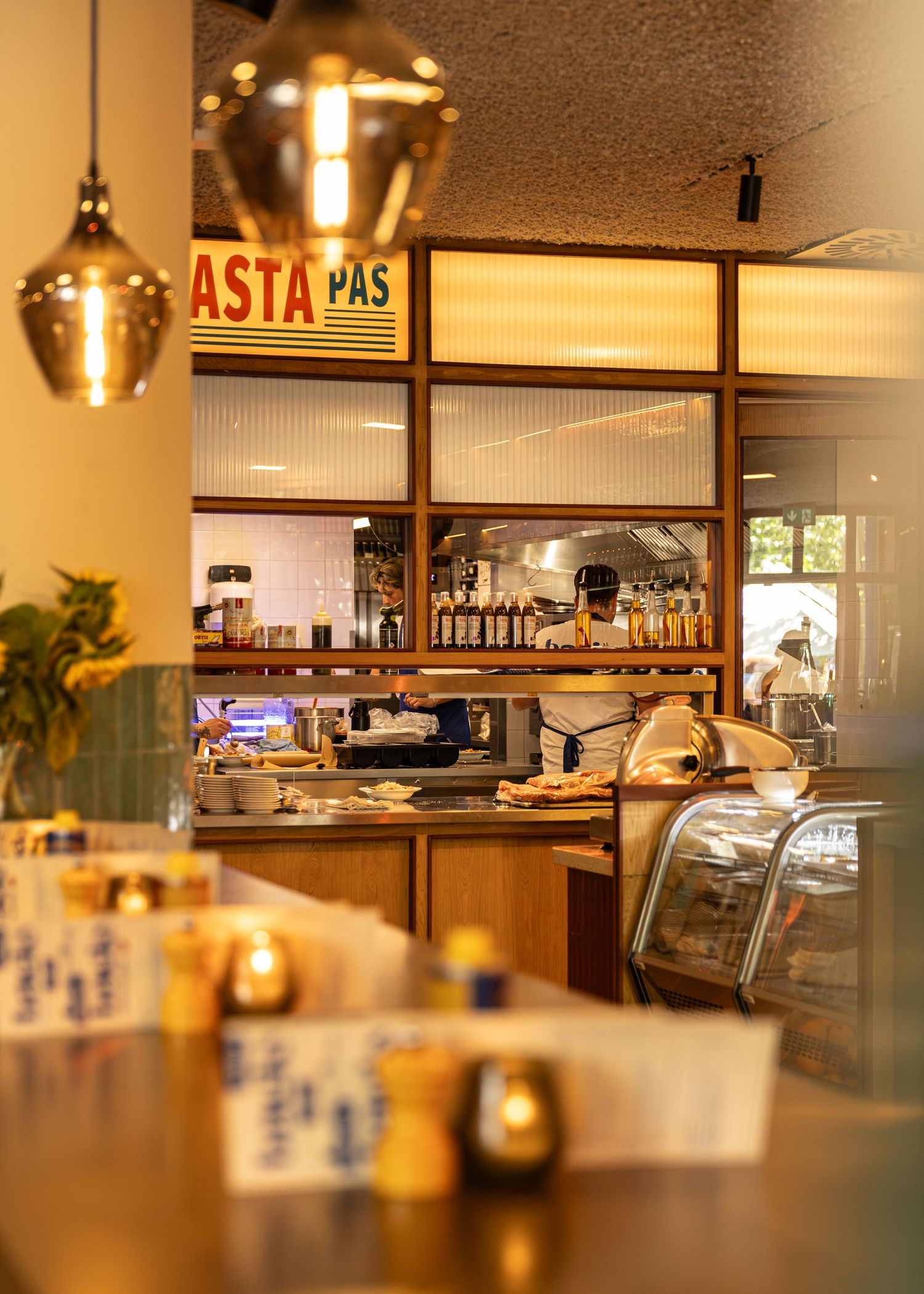 Warm Italian pasta restaurant interior with gold pendant lights, wooden counter, display case, and illuminated kitchen backdrop.