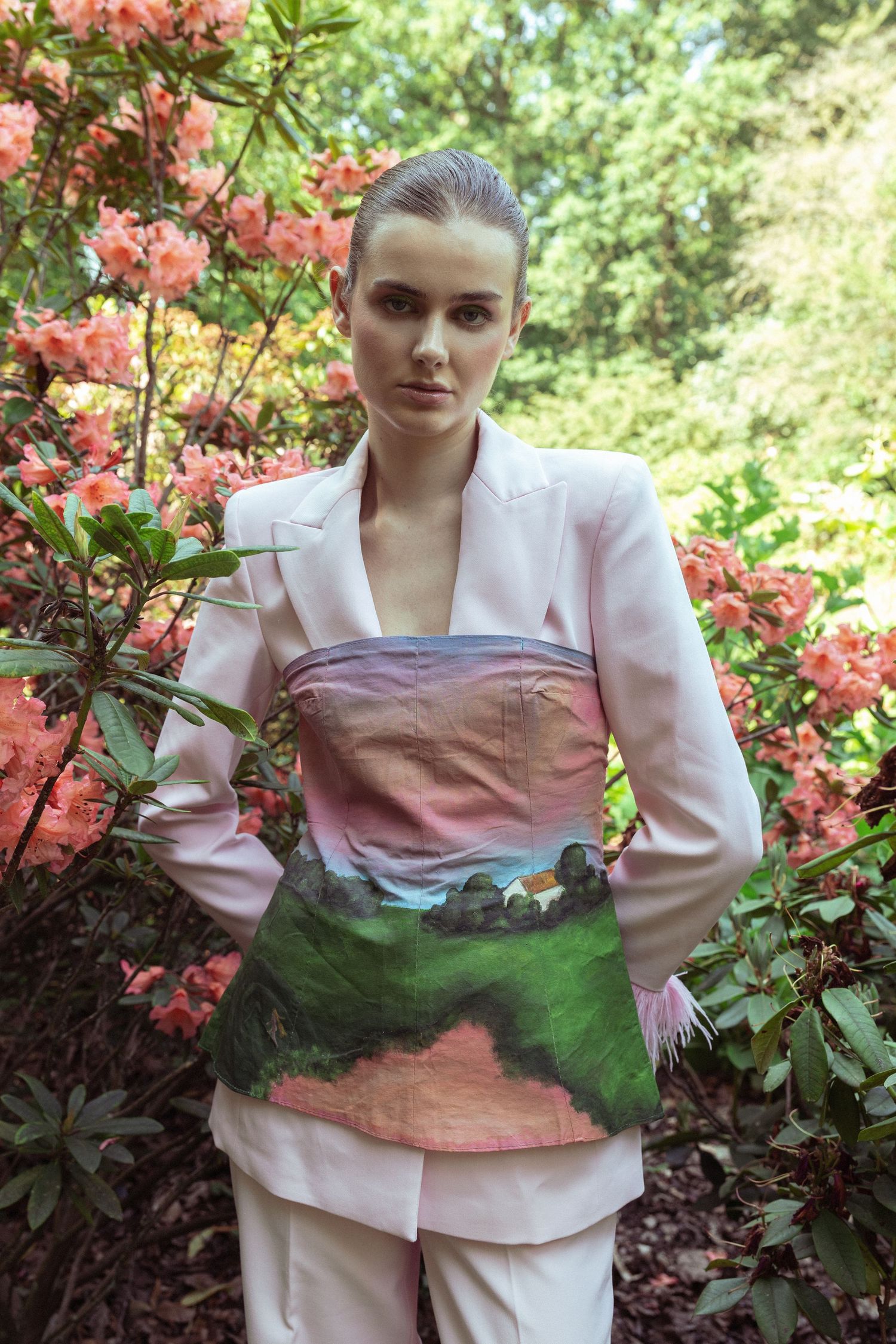 Woman in white blazer wearing a landscape-painted corset top, posing among pink flowering shrubs outdoors. fashion photography, netherlands