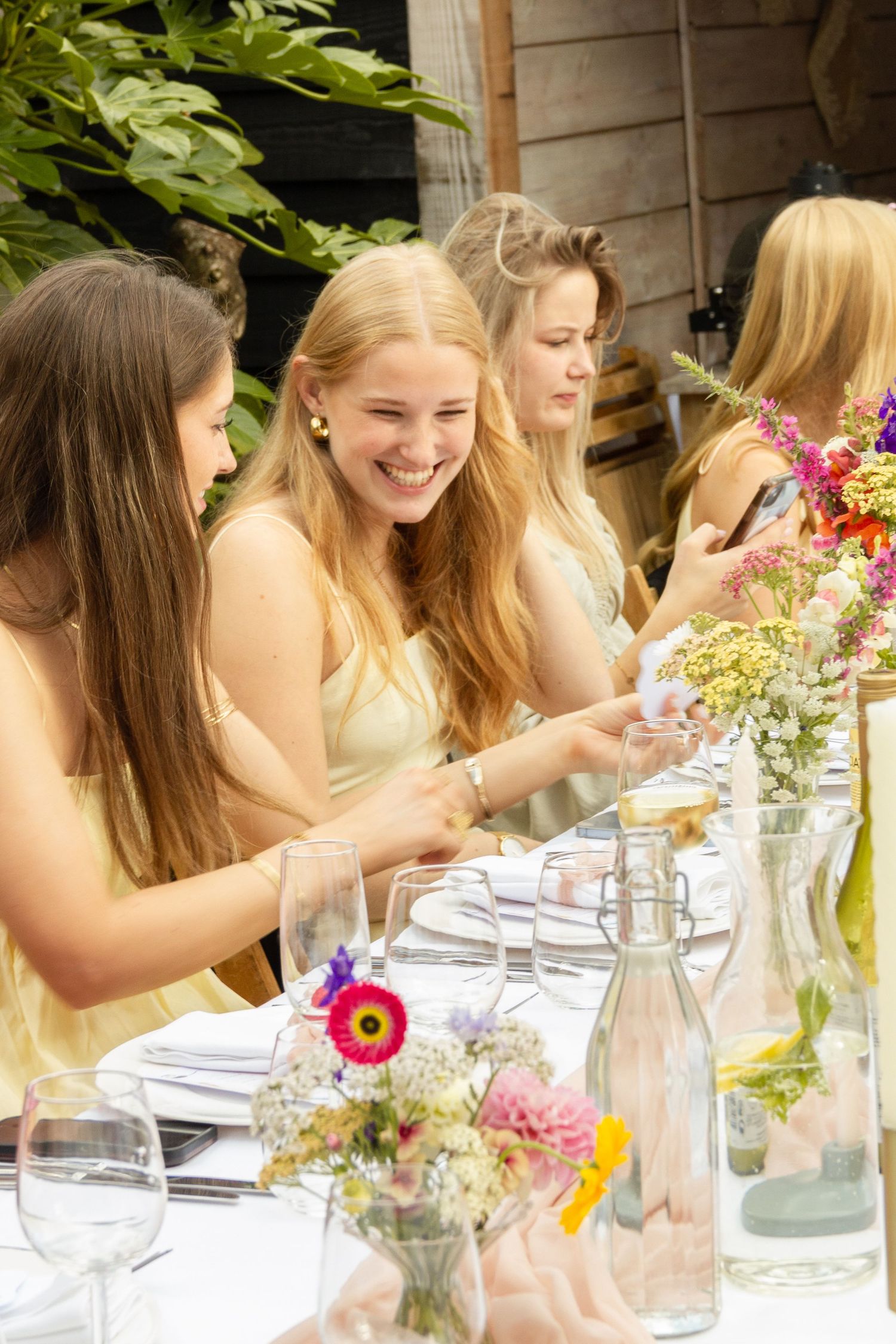 Young women in yellow dresses laughing at an outdoor garden party table decorated with colorful wildflowers. event photography, netherlands