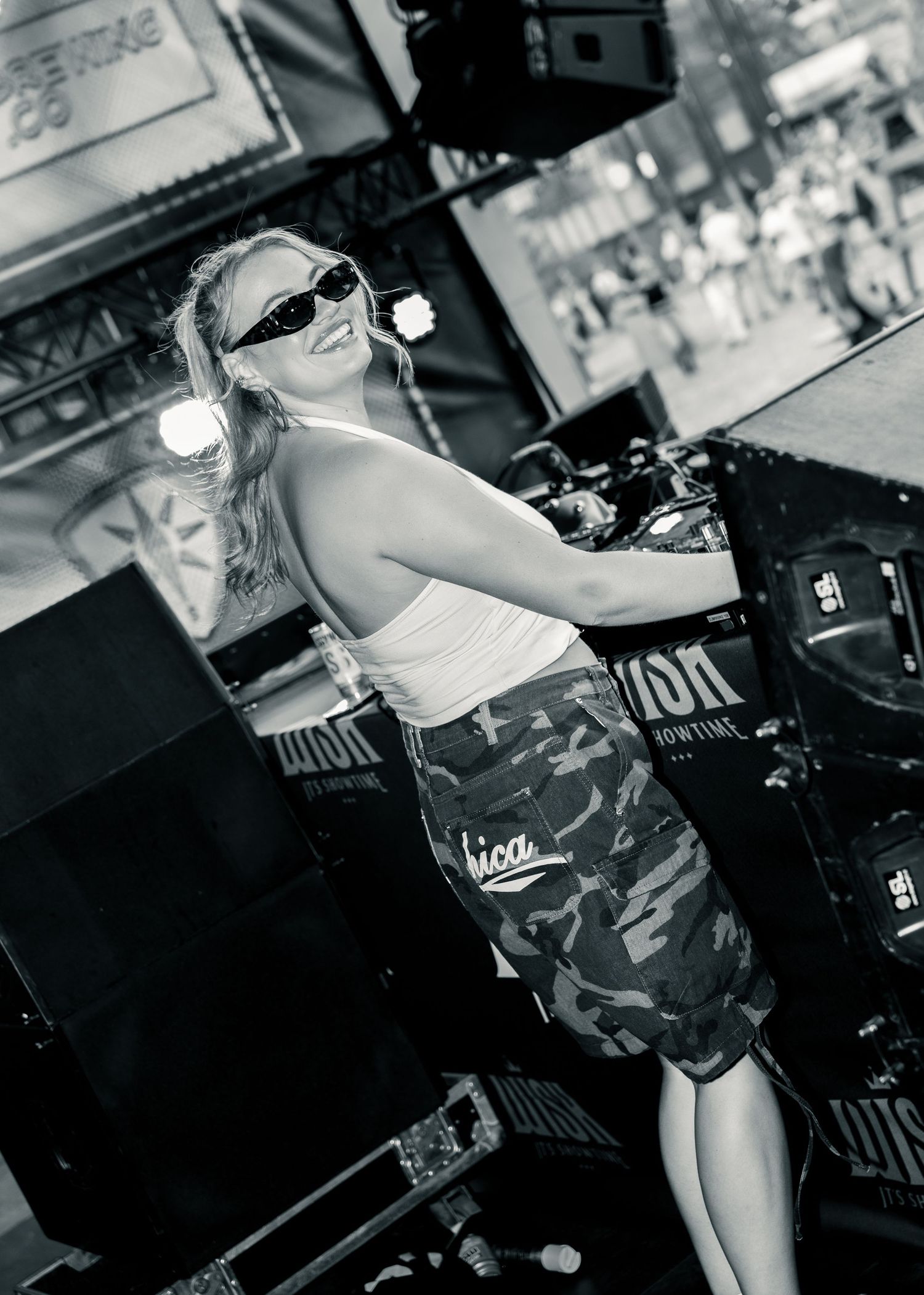 Black and white photo of a blonde woman in sunglasses, camo skirt, and tank top posing at an outdoor stage event. Festival photography,