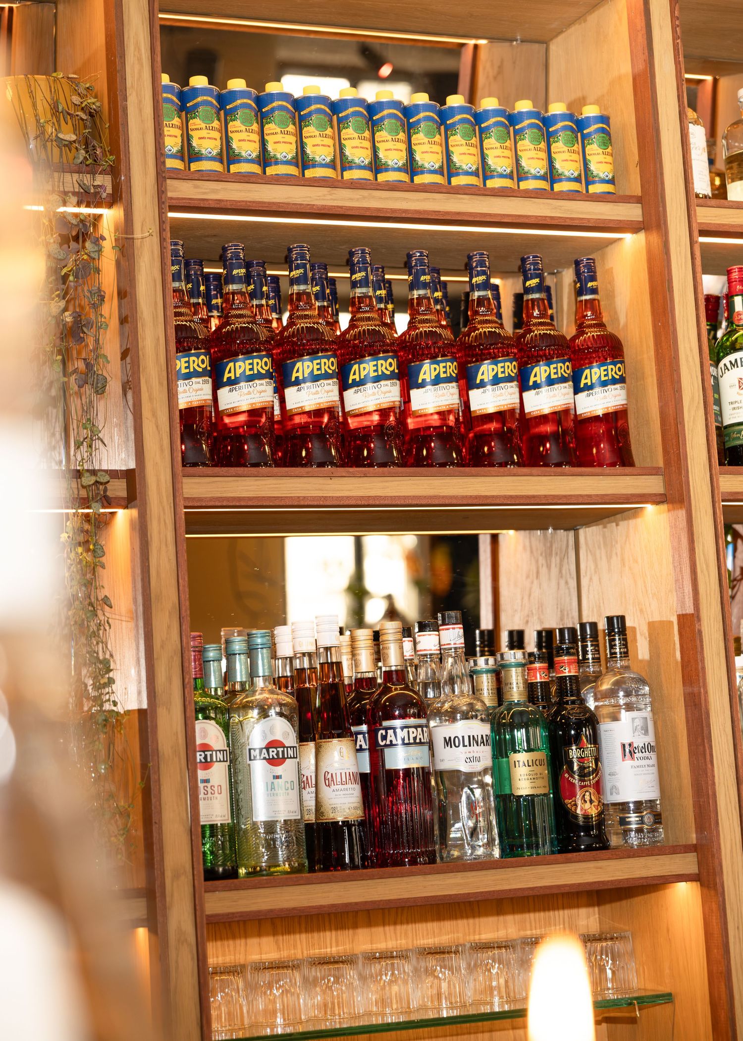 Wooden bar shelves stocked with Aperol bottles, canned drinks, and assorted liquor bottles including Martini and Campari.