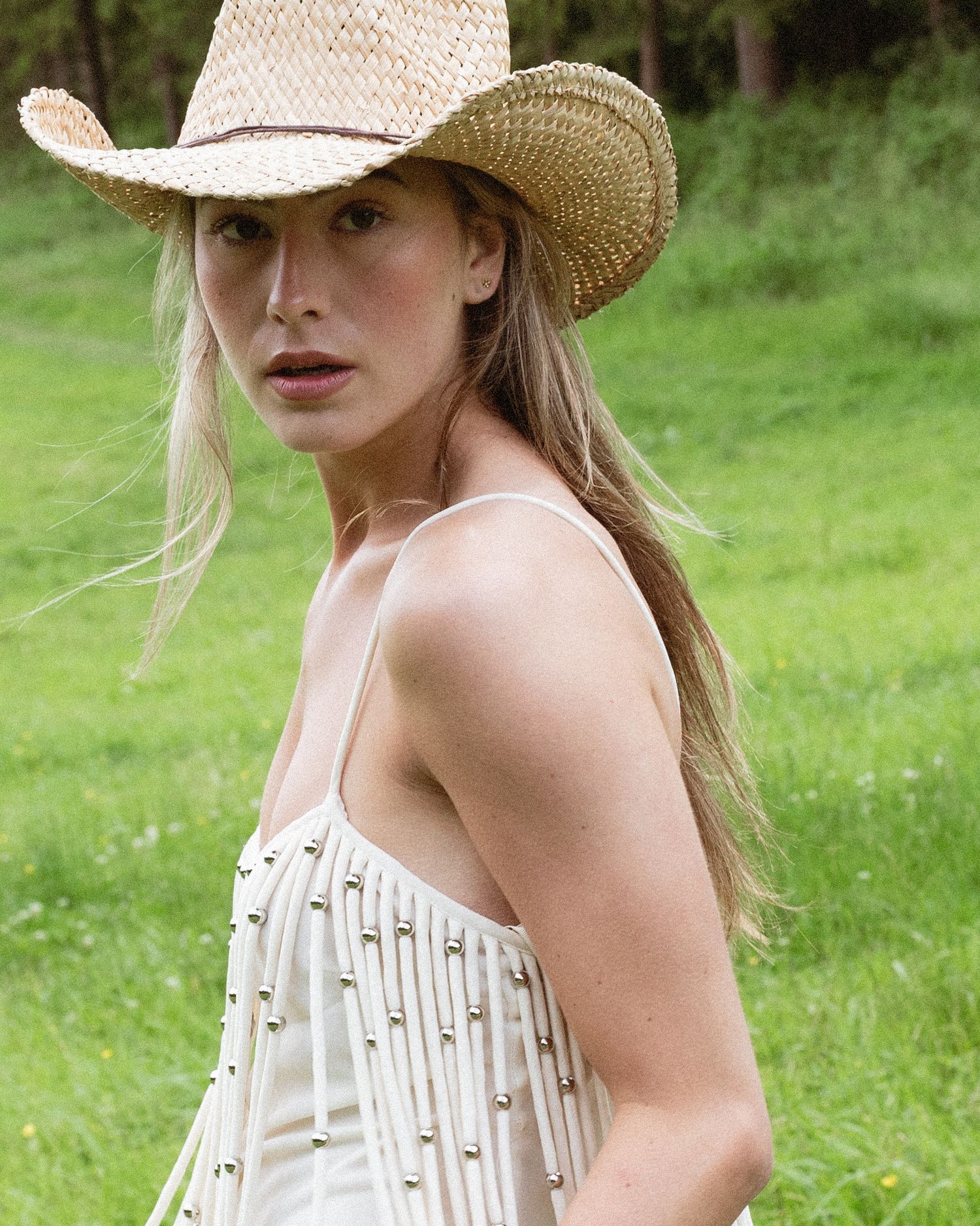 Blonde woman in a straw cowboy hat and white studded fringe top poses outdoors in a green field.  fashion photography, netherlands