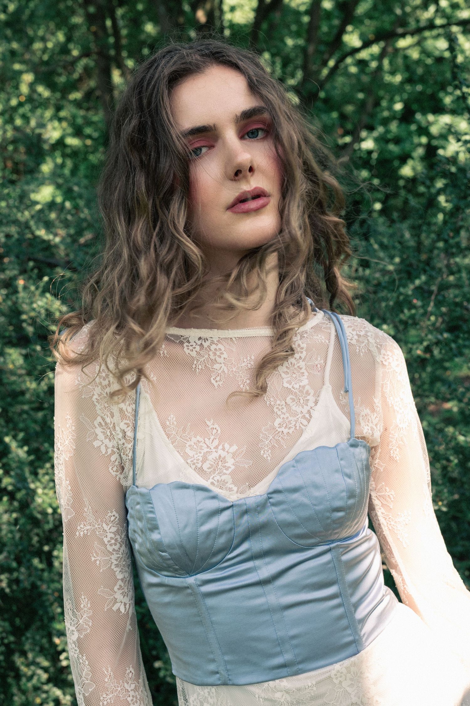 Young woman in blue satin corset and sheer lace overlay dress poses outdoors among lush green trees.  fashion photography, netherlands
