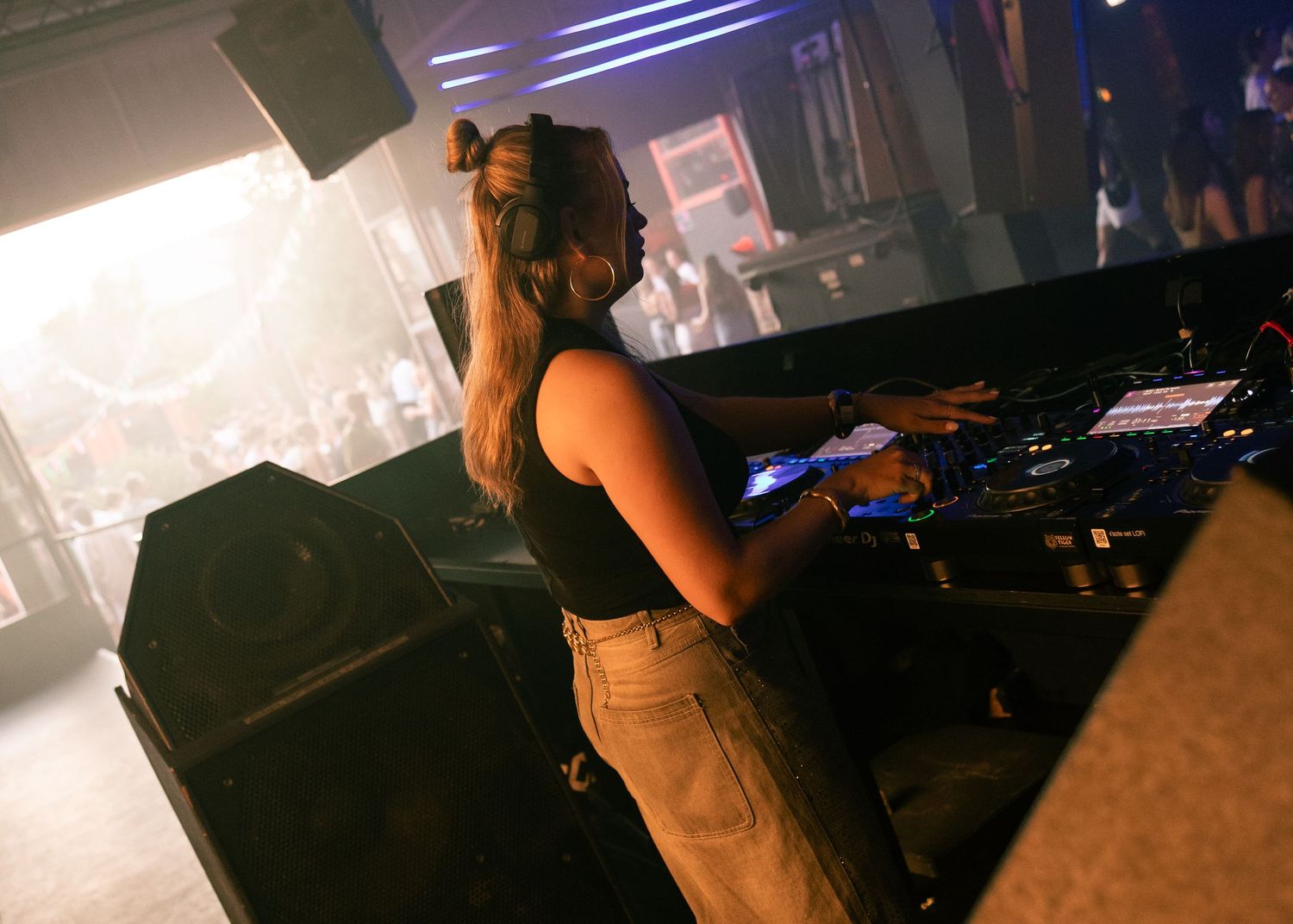 Female DJ performing at a nightclub, operating professional mixing equipment on stage in a dark venue. Festival photography,