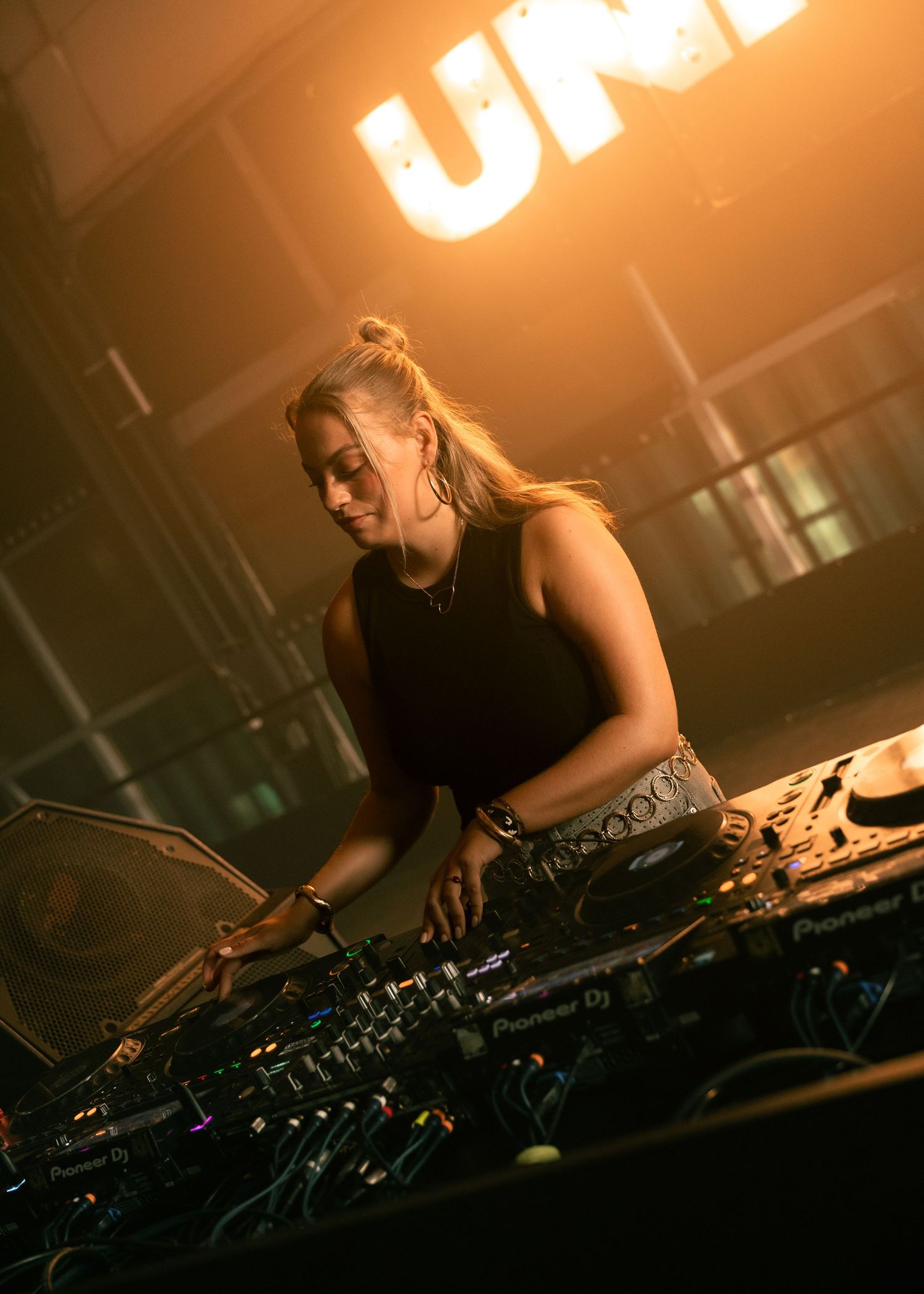 Female DJ smiling while performing on Pioneer DJ equipment under warm stage lighting at a nightclub event. Festival photography,