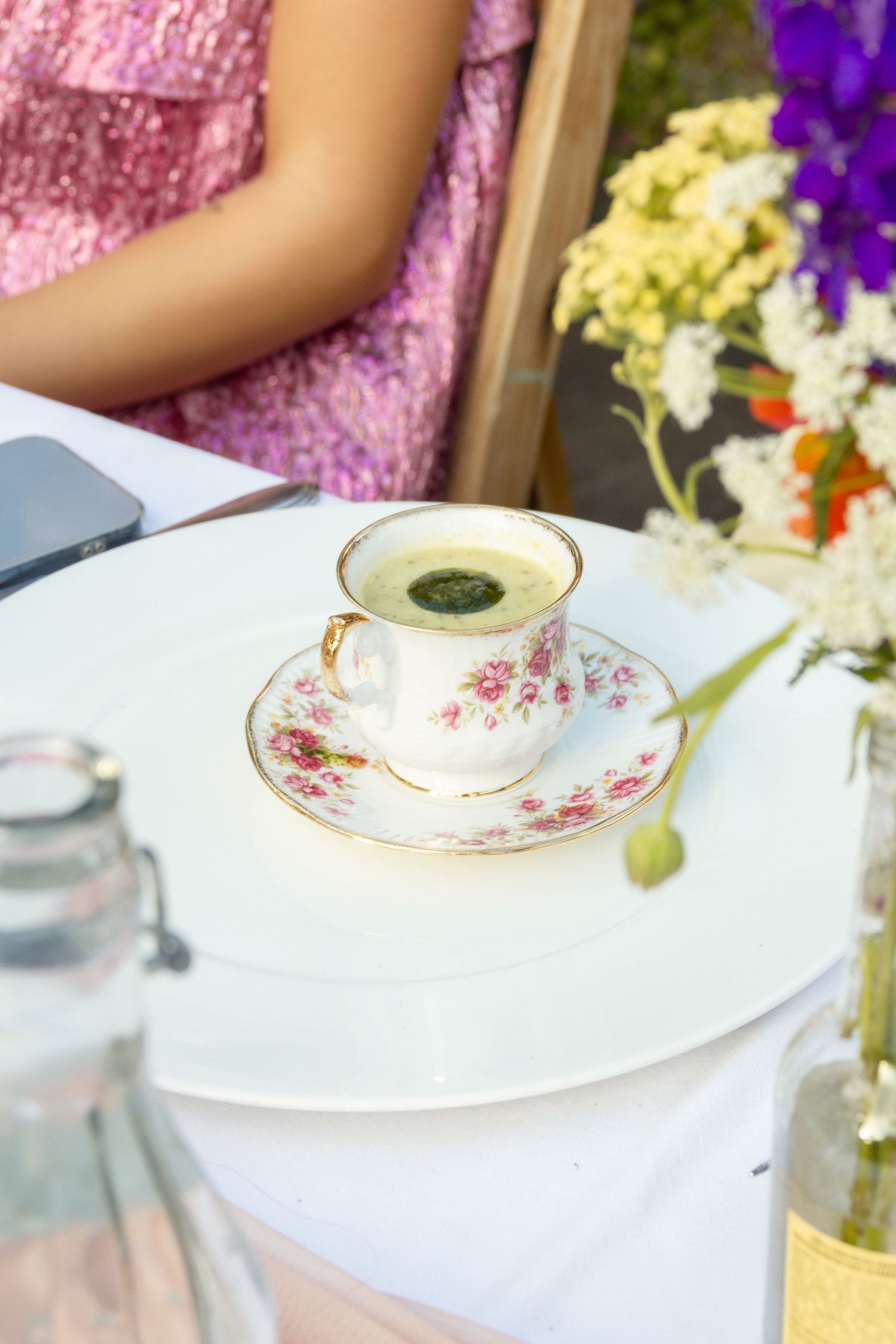 Elegant floral teacup with matcha latte on white table, woman in pink dress and flowers in background. event photography, netherlands