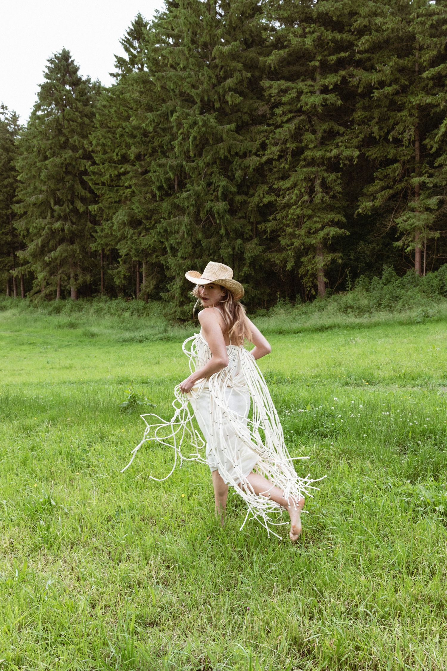 Woman in white fringe dress and straw cowboy hat walking barefoot on green grass with pine forest background.  fashion photography