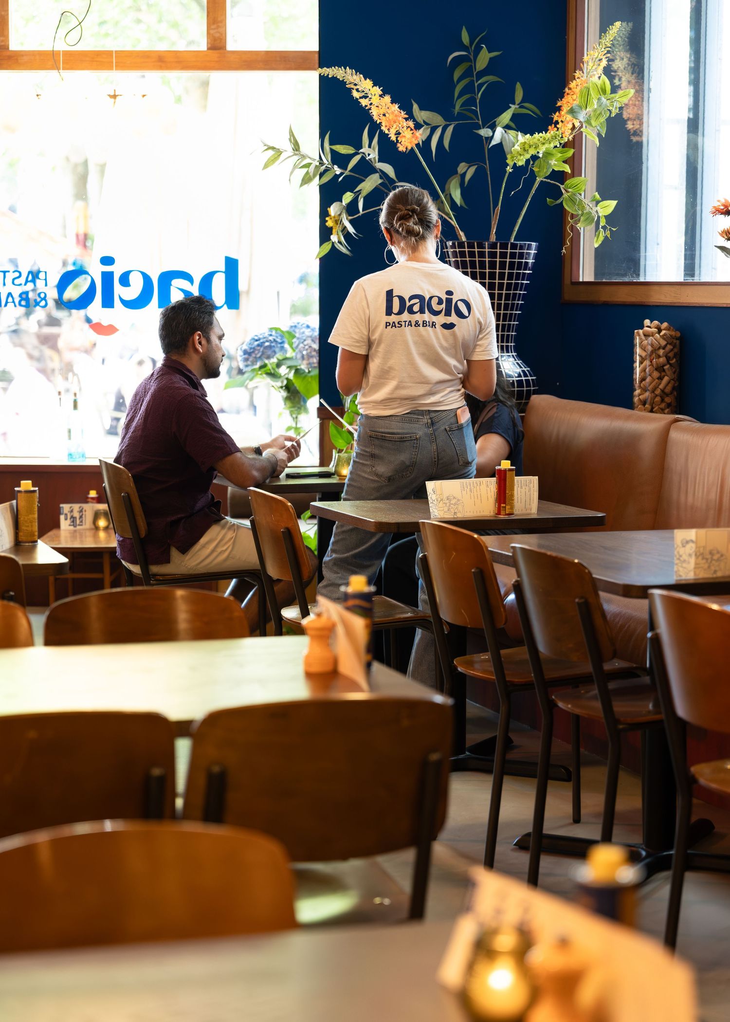 Staff member in Bacio branded shirt serving a customer inside a modern restaurant with blue walls and wooden furniture.