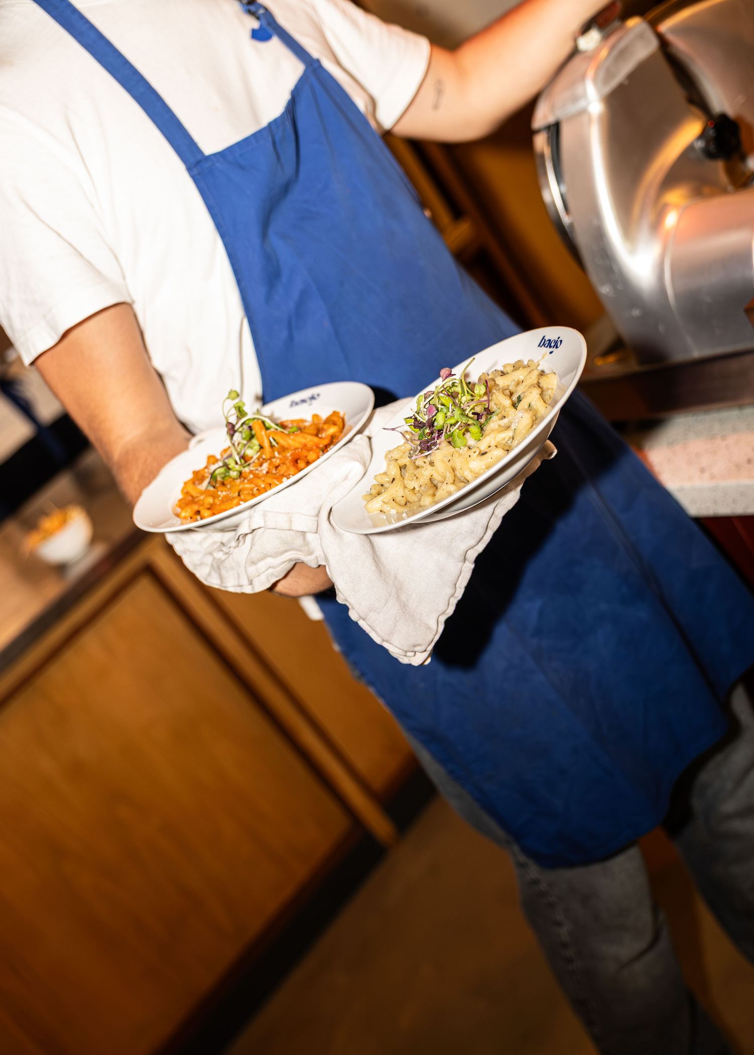 A server in a blue apron holds two plates of colorful pasta dishes in a warm restaurant kitchen.