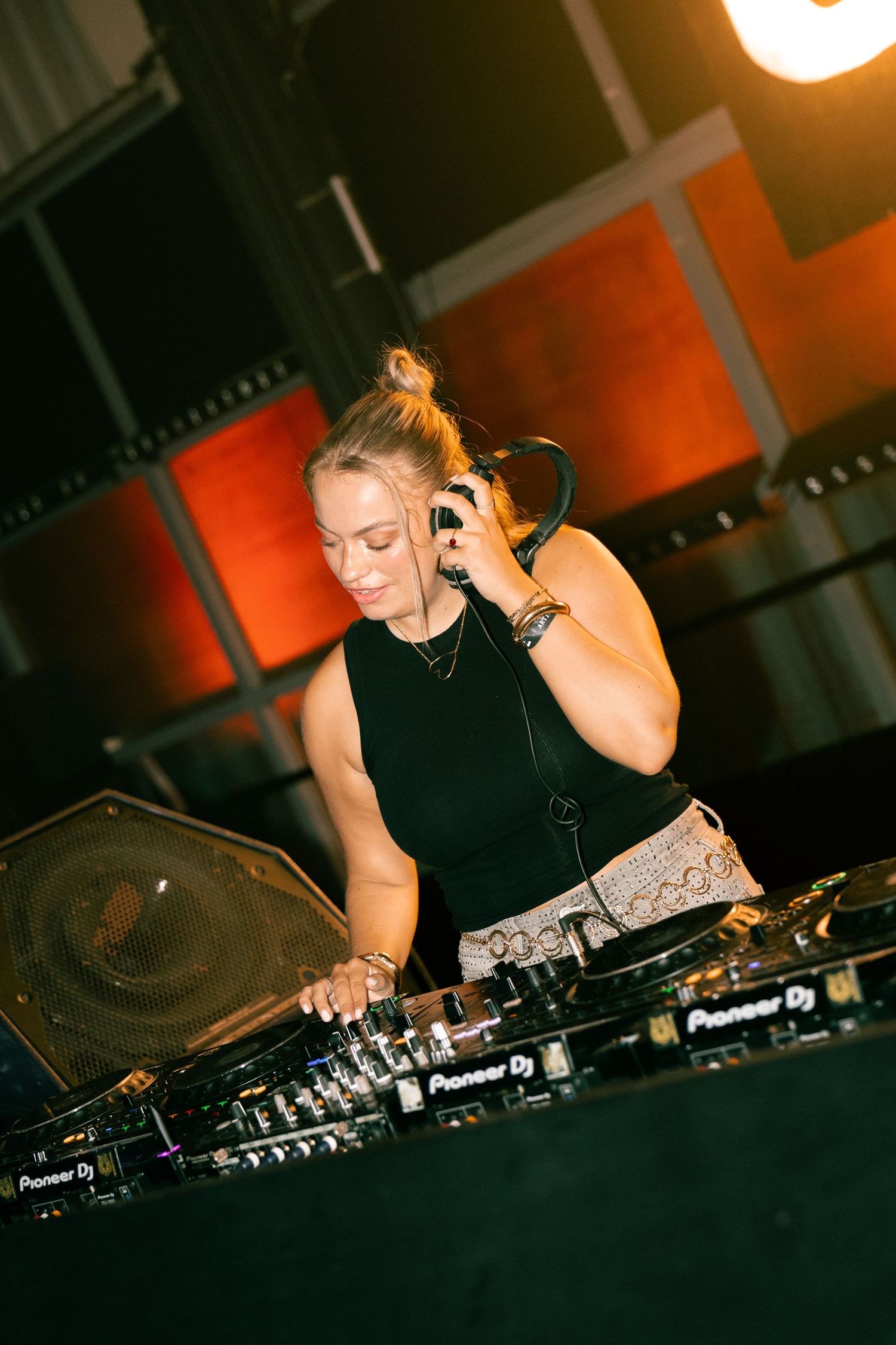 Female DJ mixing tracks on Pioneer DJ equipment while wearing headphones in a dimly lit venue with orange lighting. Festival photography,