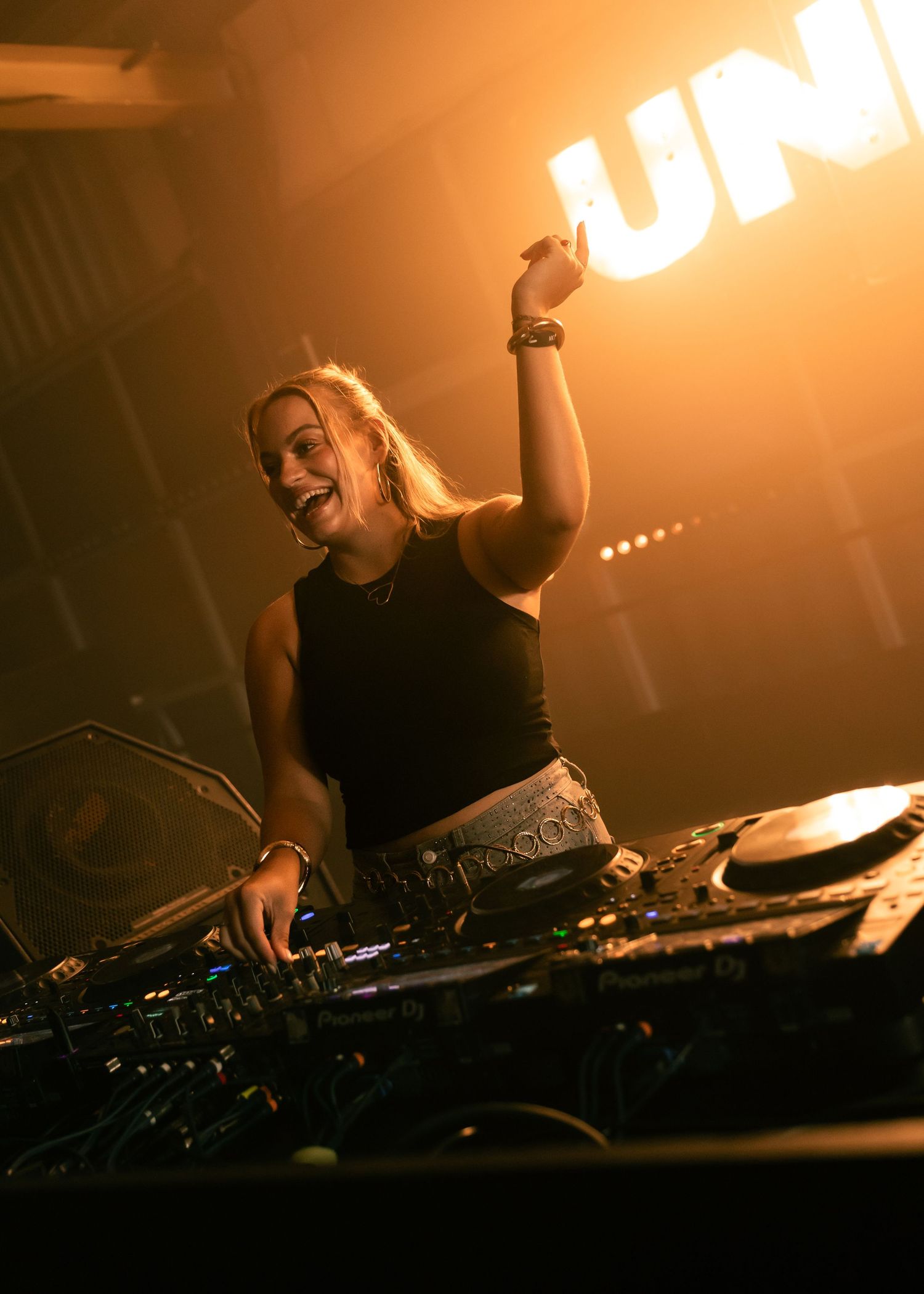 Female DJ performing energetically at a club, hands raised behind DJ decks under warm golden stage lighting. Festival photography,