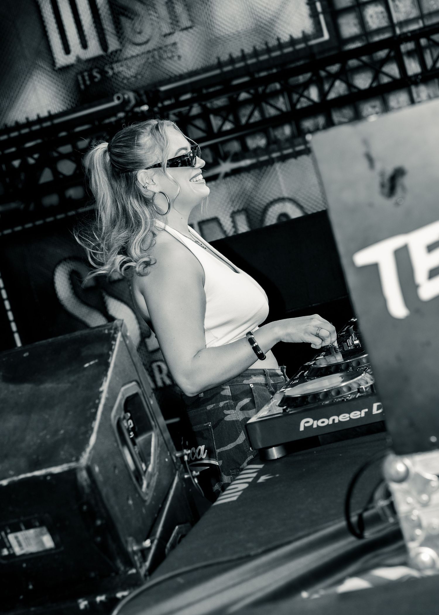 Female DJ with blonde ponytail and sunglasses mixing on Pioneer DJ equipment at an outdoor music event. Festival photography,