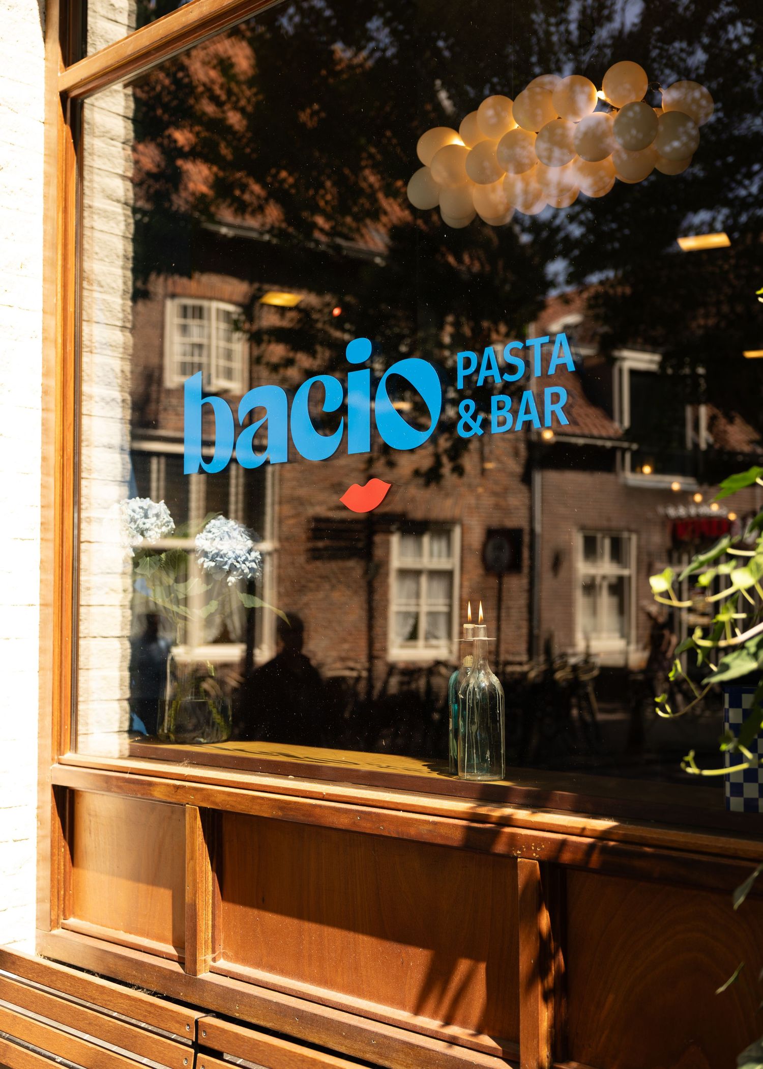 Bacio Pasta & Bar restaurant storefront window with blue logo, balloons, and flower vases reflecting a sunny street scene.