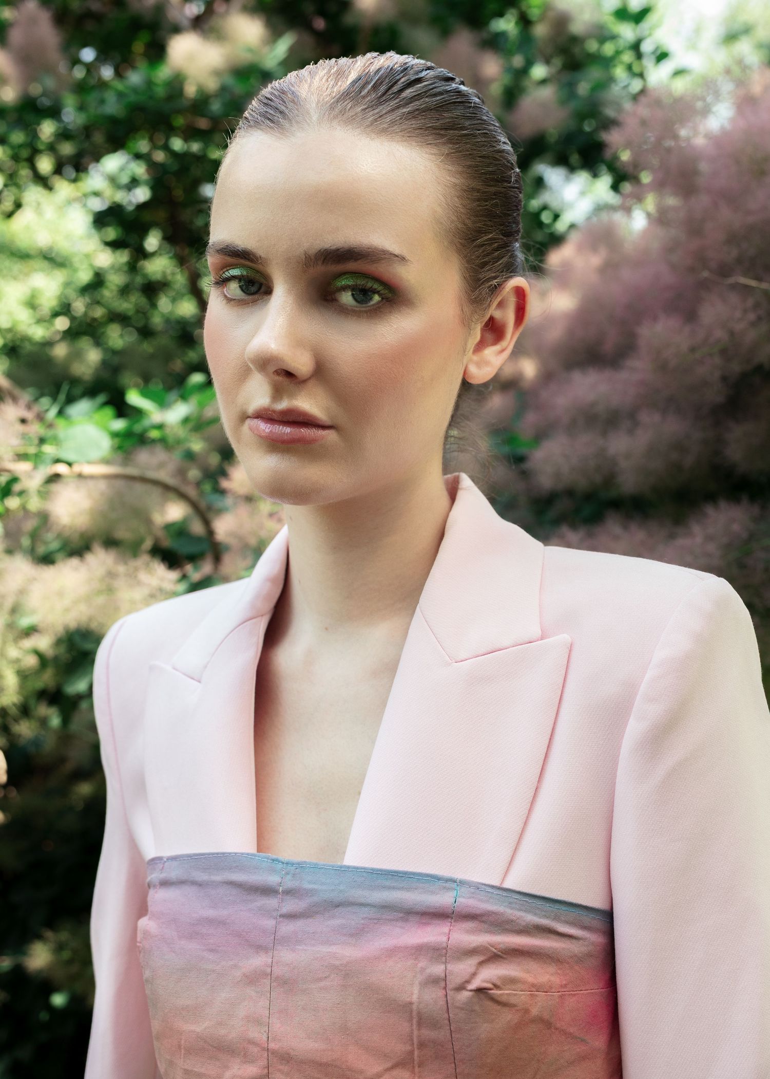 A woman in a pink blazer with sleek hair and green eyeshadow poses outdoors among lush pink-blossomed trees. fashion photography,