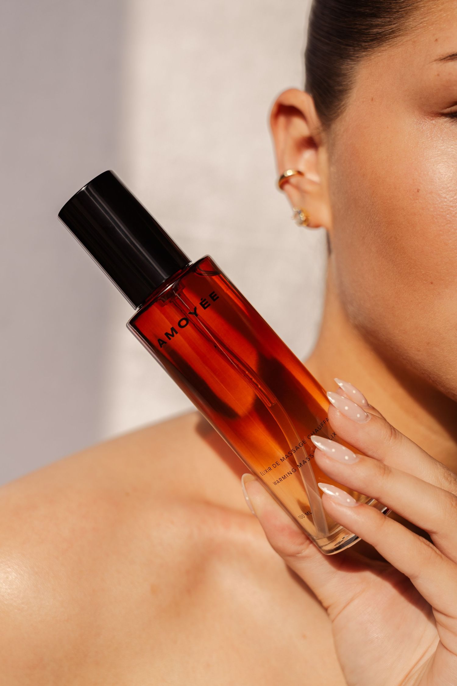 Woman holding oil bottle perfume bottle with black cap, showcasing glowing skin and French manicure nails.  fashion photography, netherlands