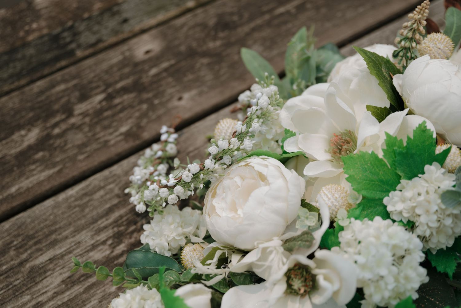 White peonies and baby's breath arranged with green foliage on rustic wooden planks.