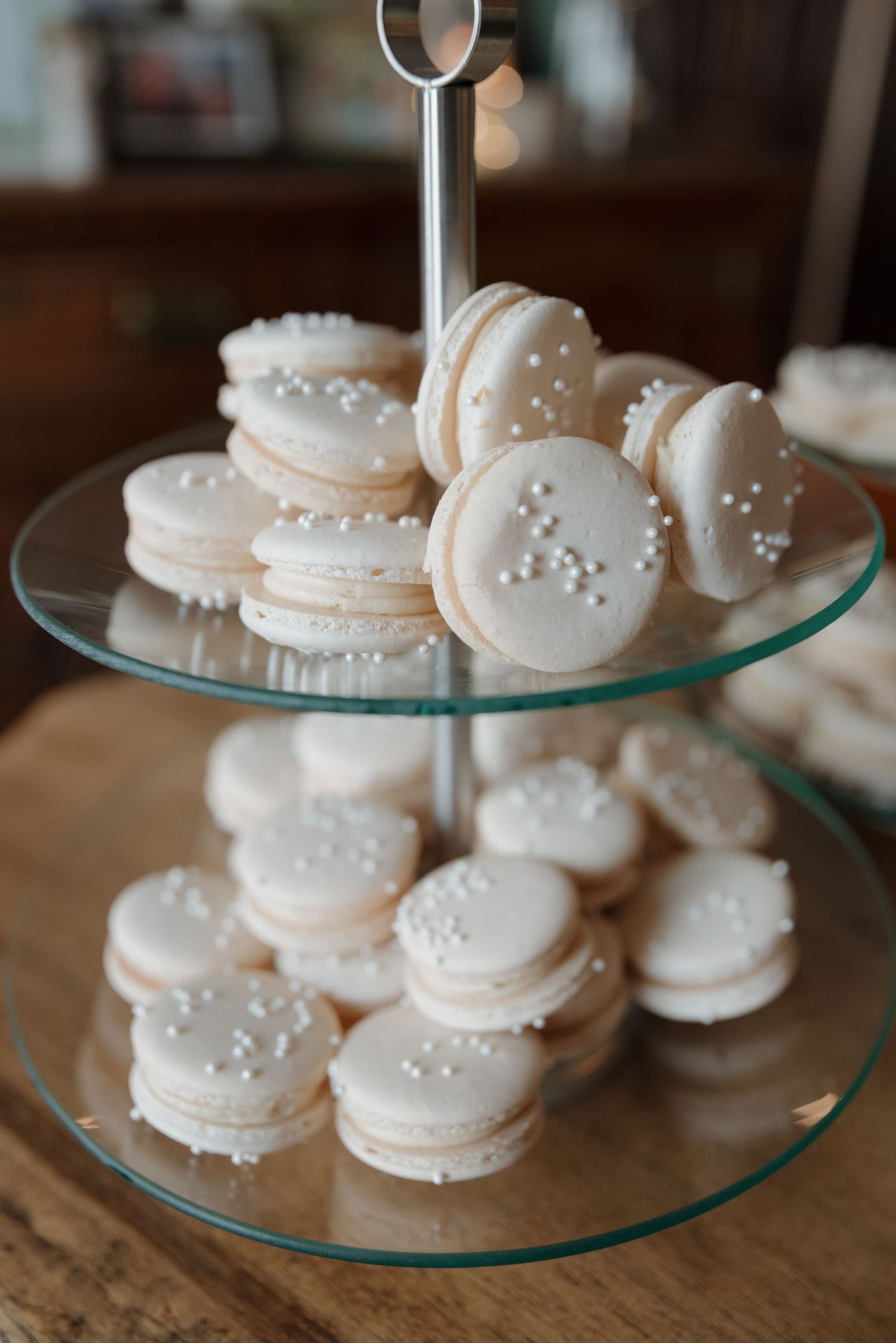White macarons decorated with pearl sprinkles displayed on a tiered glass serving stand.