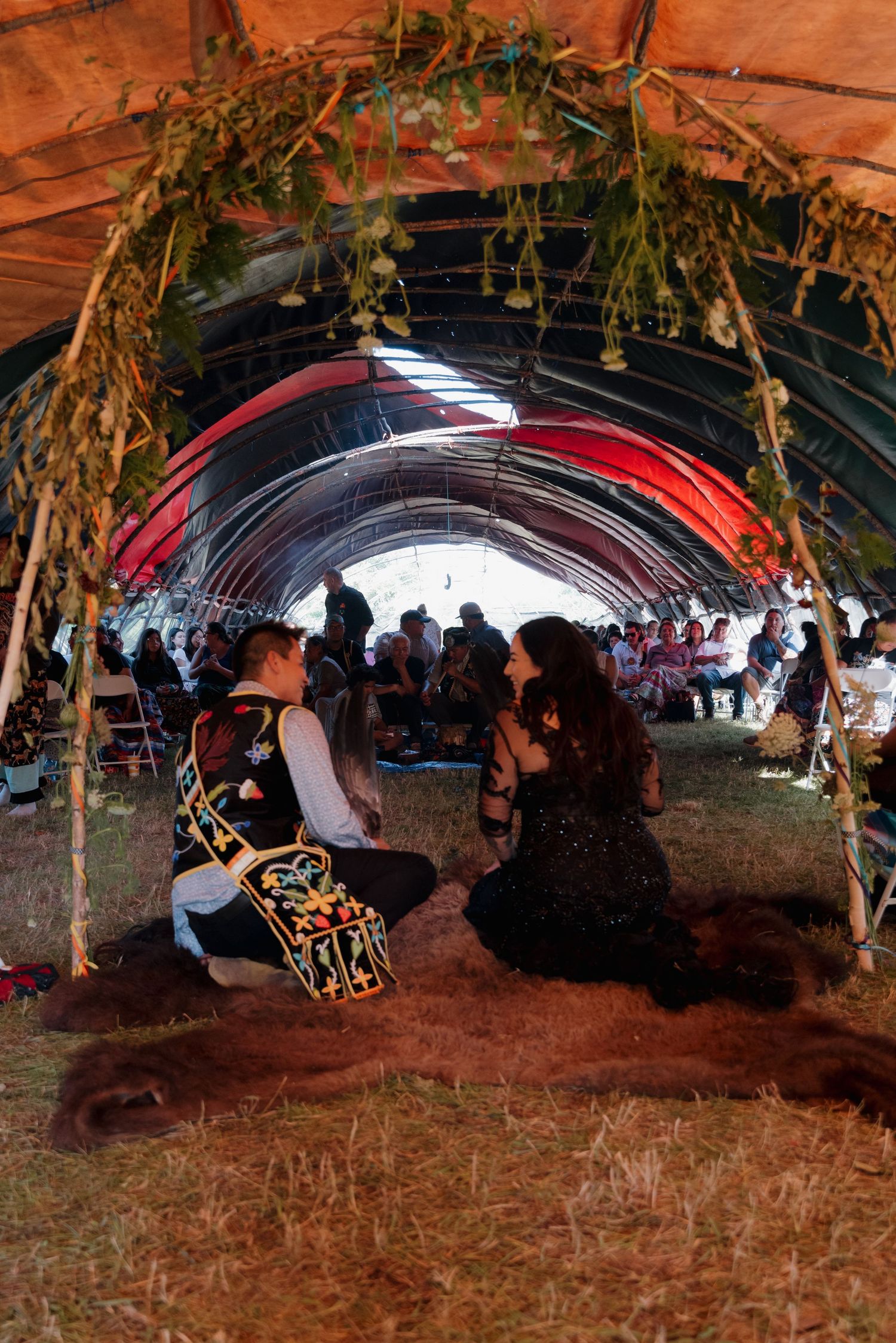 Couple kneels together under floral arch on animal hide during intimate Native American wedding ceremony inside ceremonial tent.