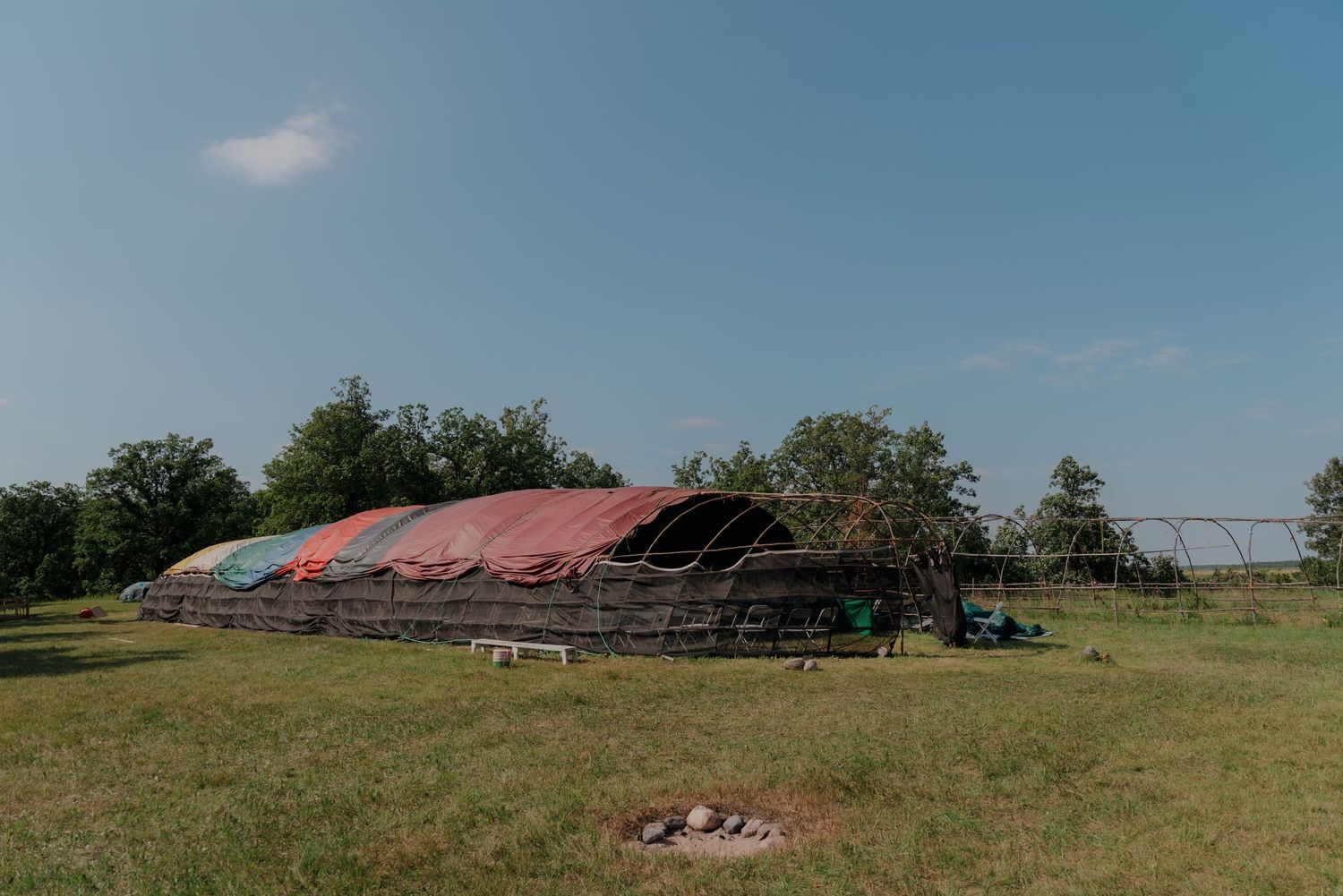 A large colorful tarp-covered greenhouse structure on a green field with trees in the background under a blue sky.