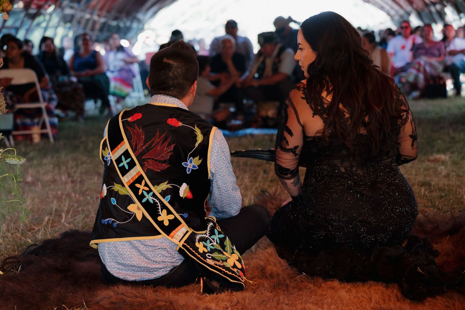 Couple in traditional Native American regalia sitting on animal fur during an outdoor cultural ceremony under a hide arbor.