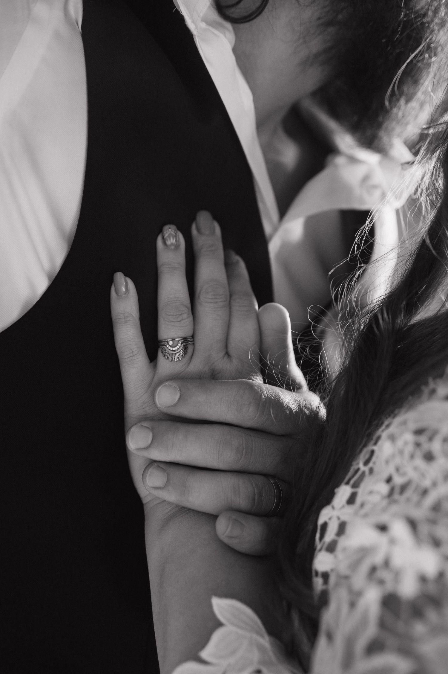 Black and white close-up of bride's hand with diamond ring resting on groom's arm in lace sleeve.