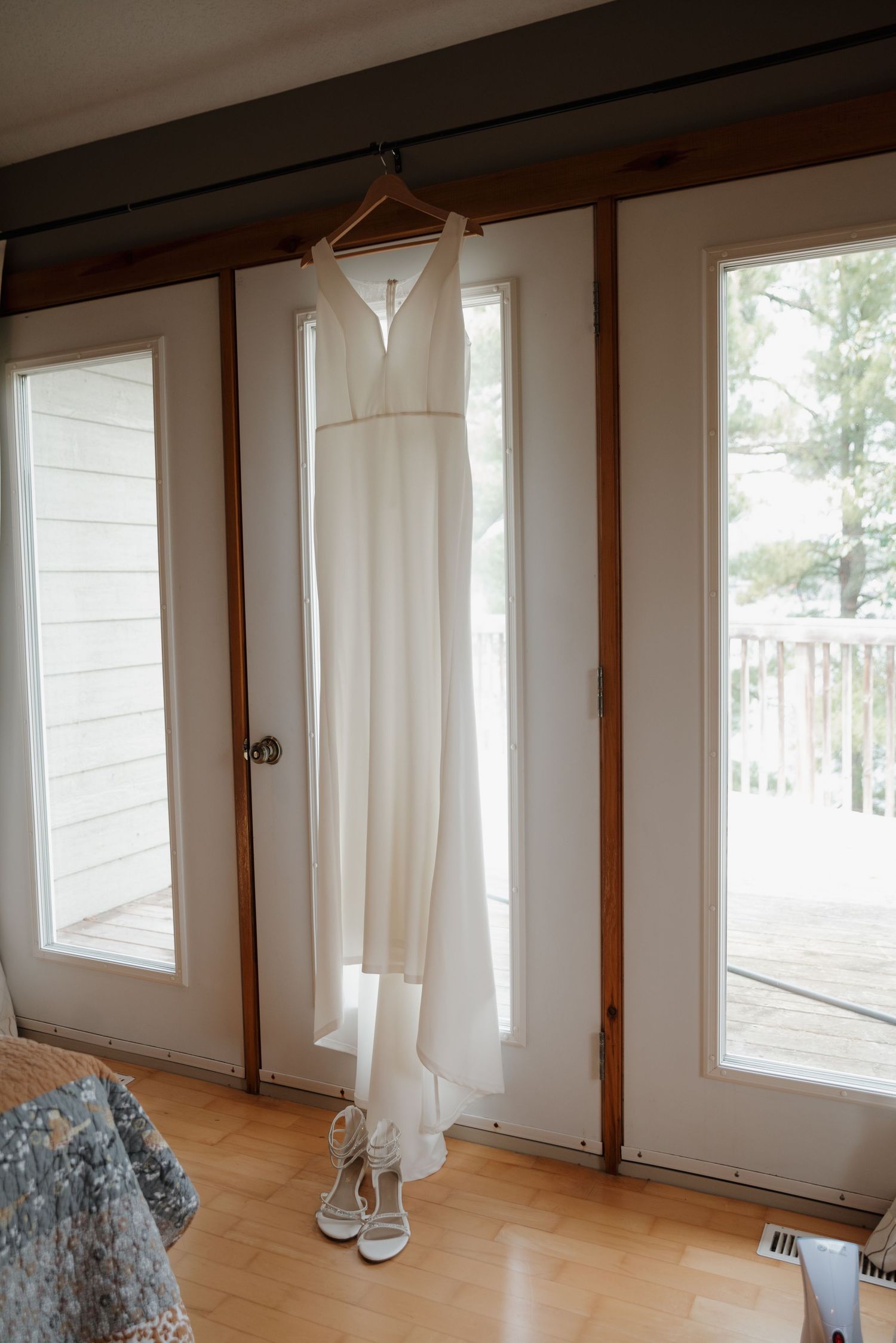A simple white wedding dress hangs by a window with white shoes beneath it.