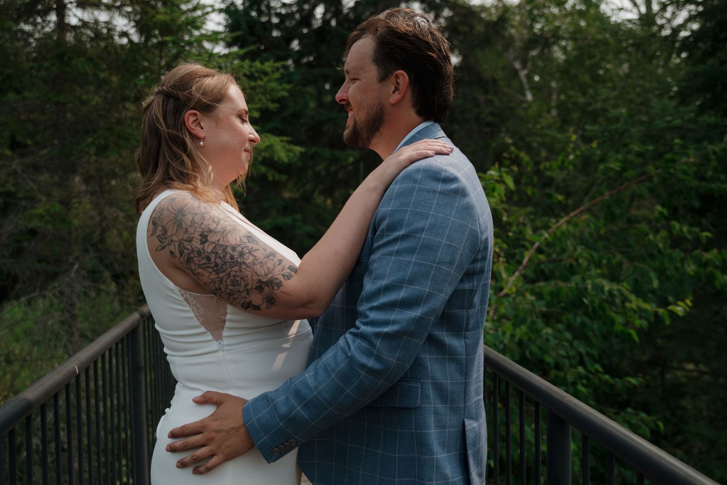 A couple embraces on a deck railing surrounded by green trees during a wedding celebration.