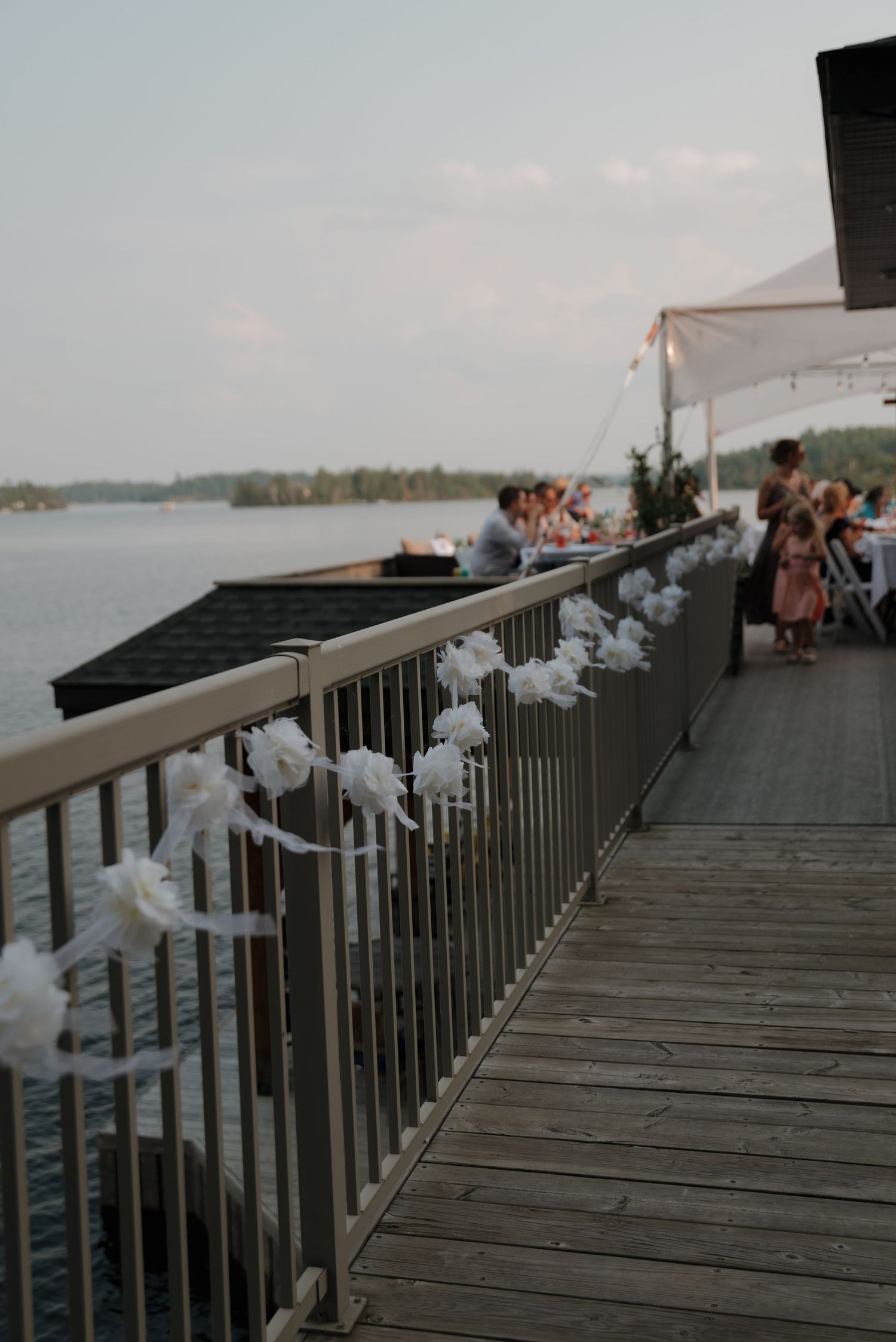 White floral decorations adorn a wooden dock railing overlooking a lake at sunset.