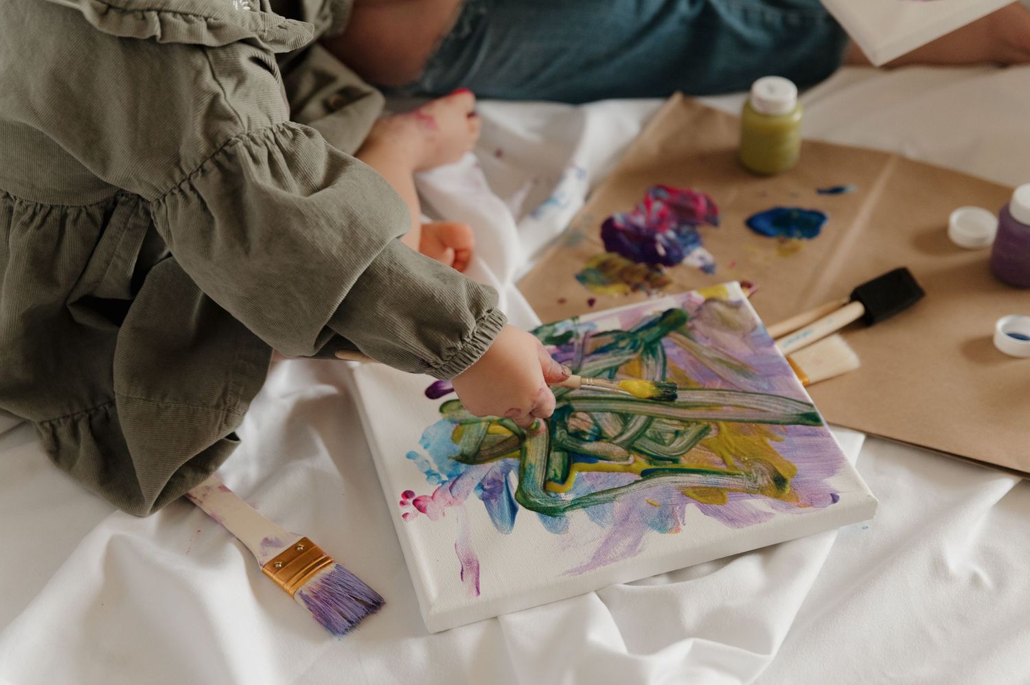 Child painting colorful artwork on canvas with brushes and paint on white cloth.