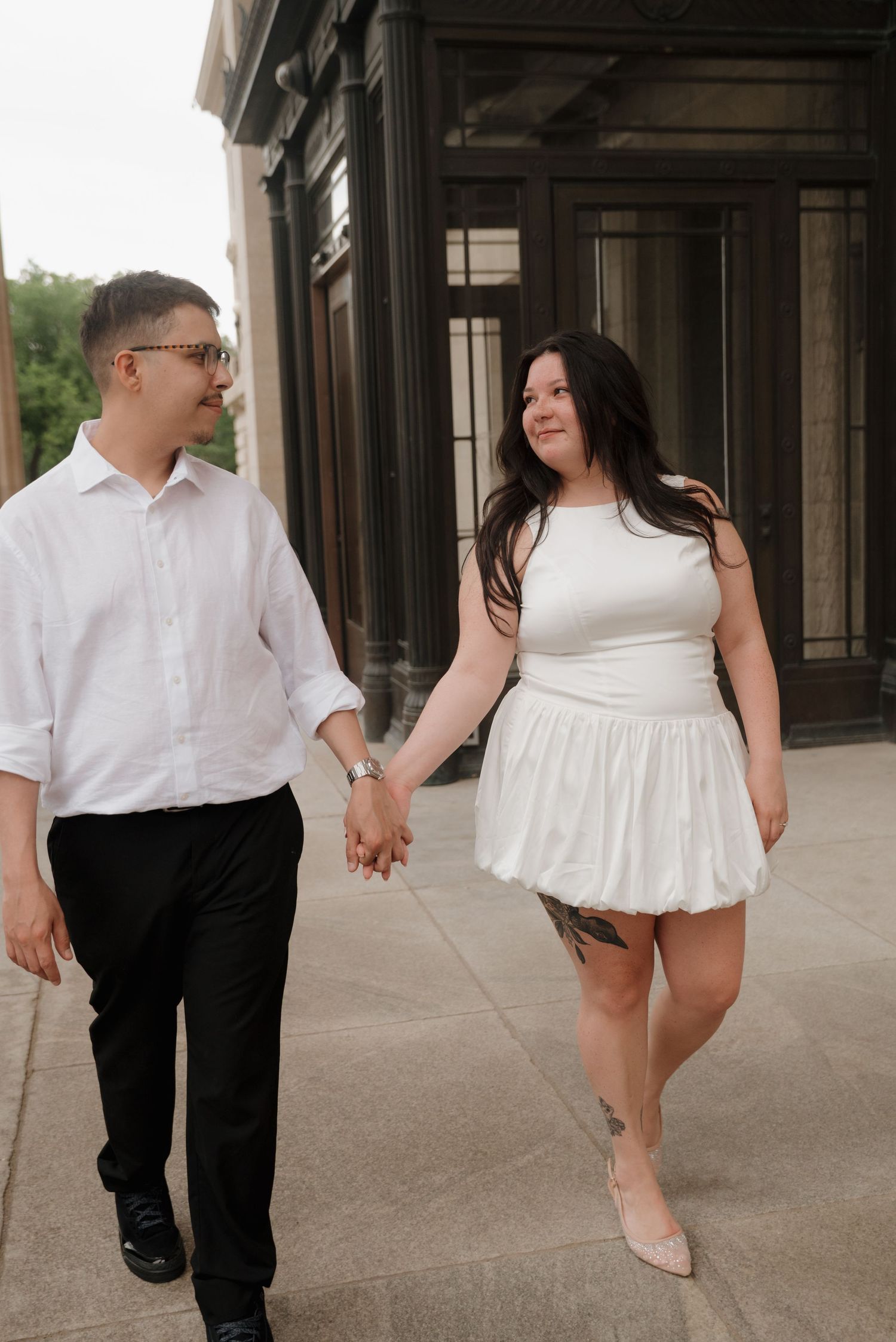 A couple holding hands while walking together outside a building, with one person in white and one in dark attire.