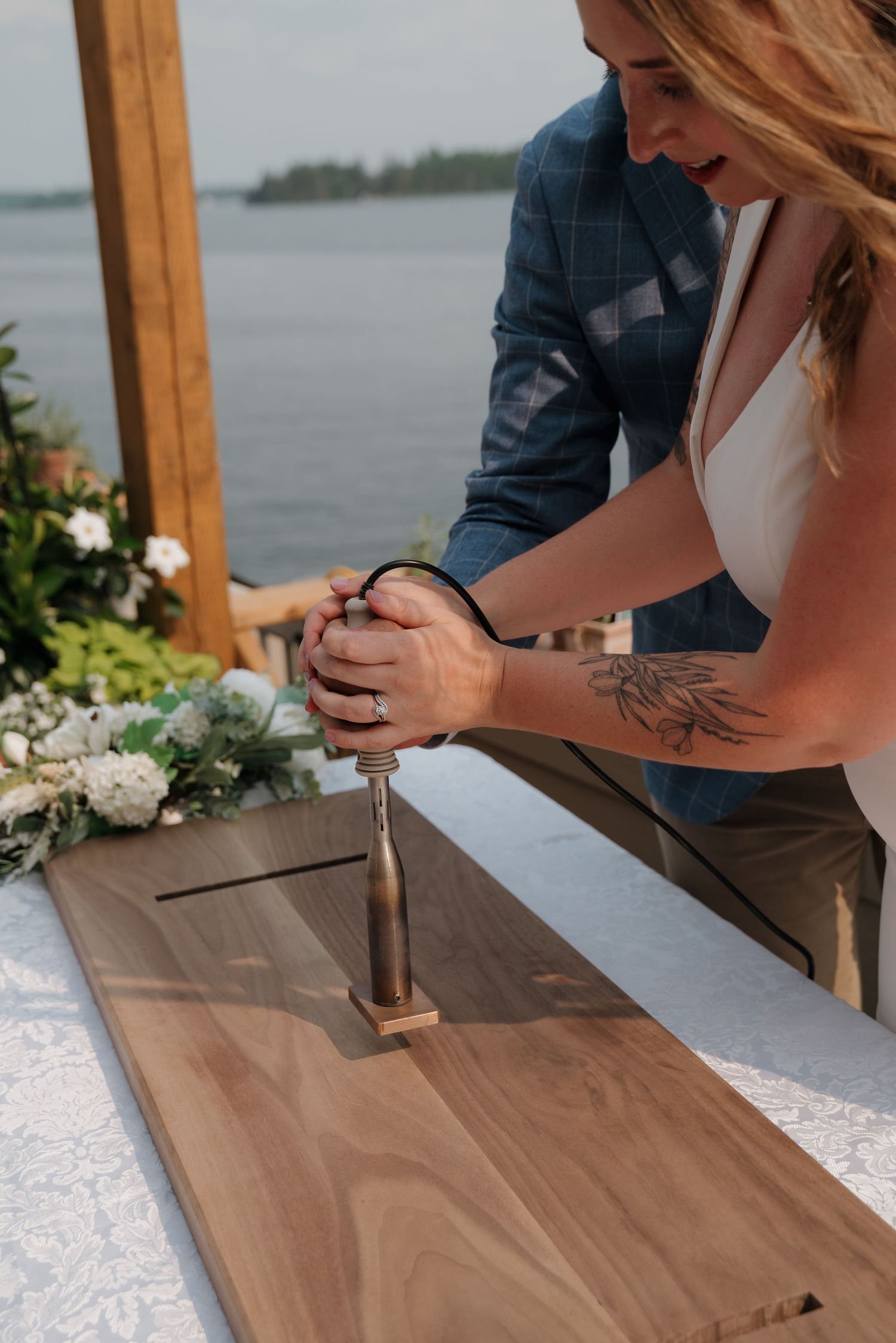 Two people signing a wooden board at a waterfront wedding ceremony with white floral decor.