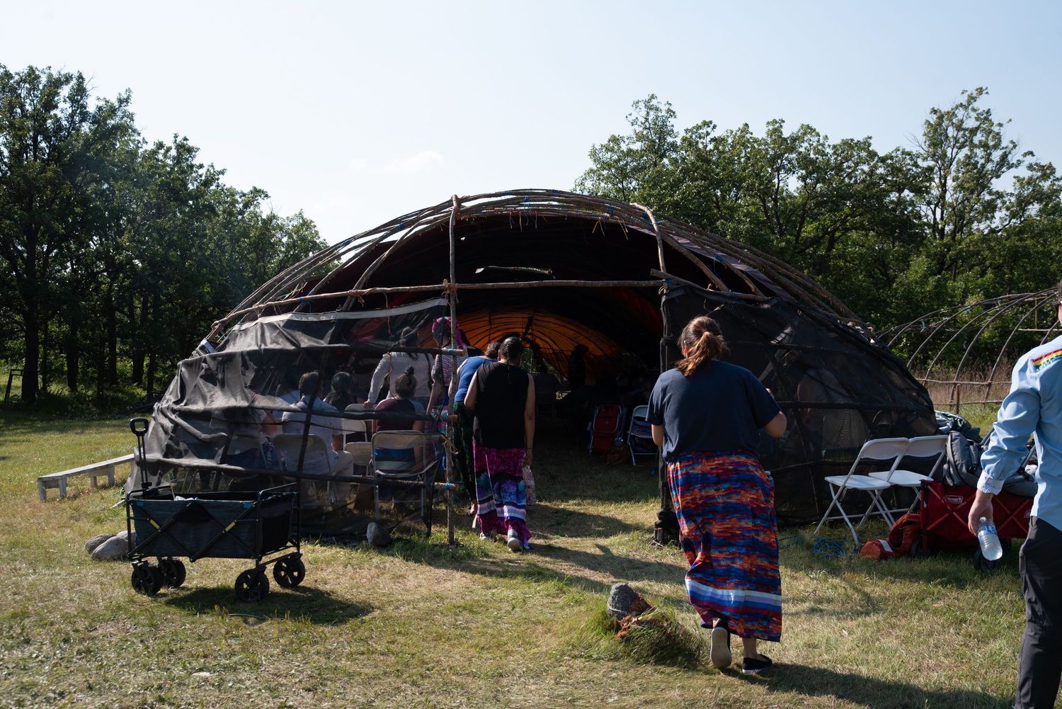 Traditional Native American dome-shaped ceremonial structure with people in colorful regalia gathered outside on a sunny day.