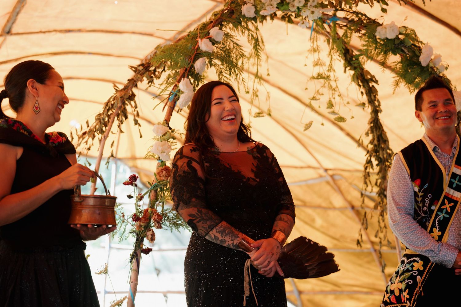 Three people laughing joyfully under a floral arch at an outdoor wedding ceremony.